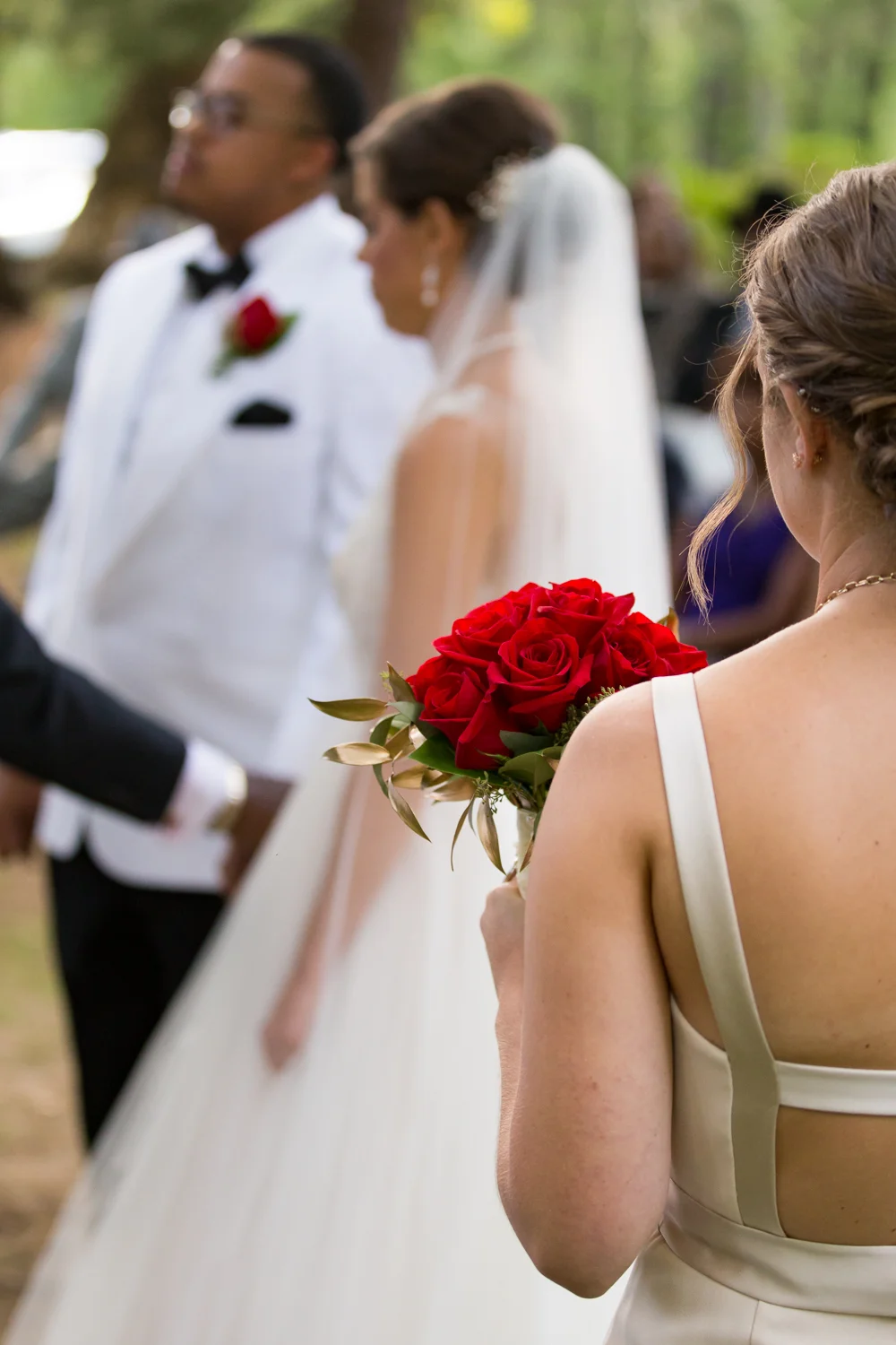 Photo of bridesmaid holding red rose bouquet with bride and groom in the background