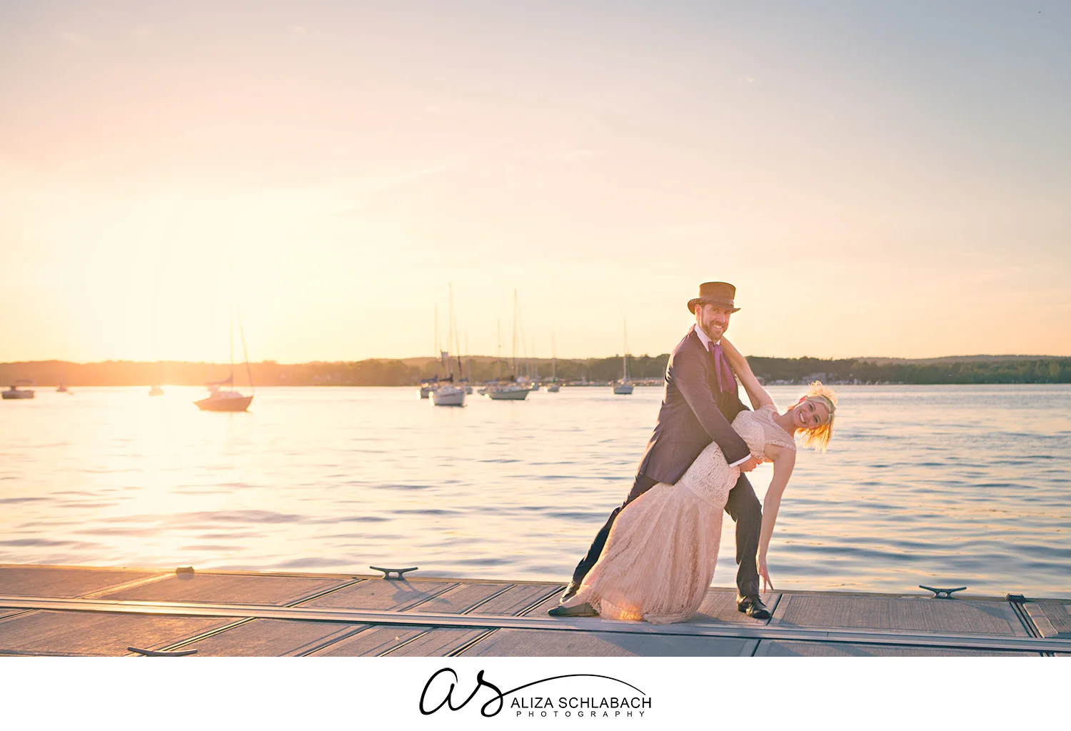 Photo of Groom in a tophat dipping his bride at sunset over the bay with sailboats