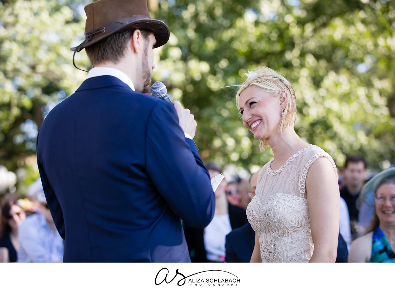 Photo of a blonde bride smiling at her groom while he speaks his vows into a microphone