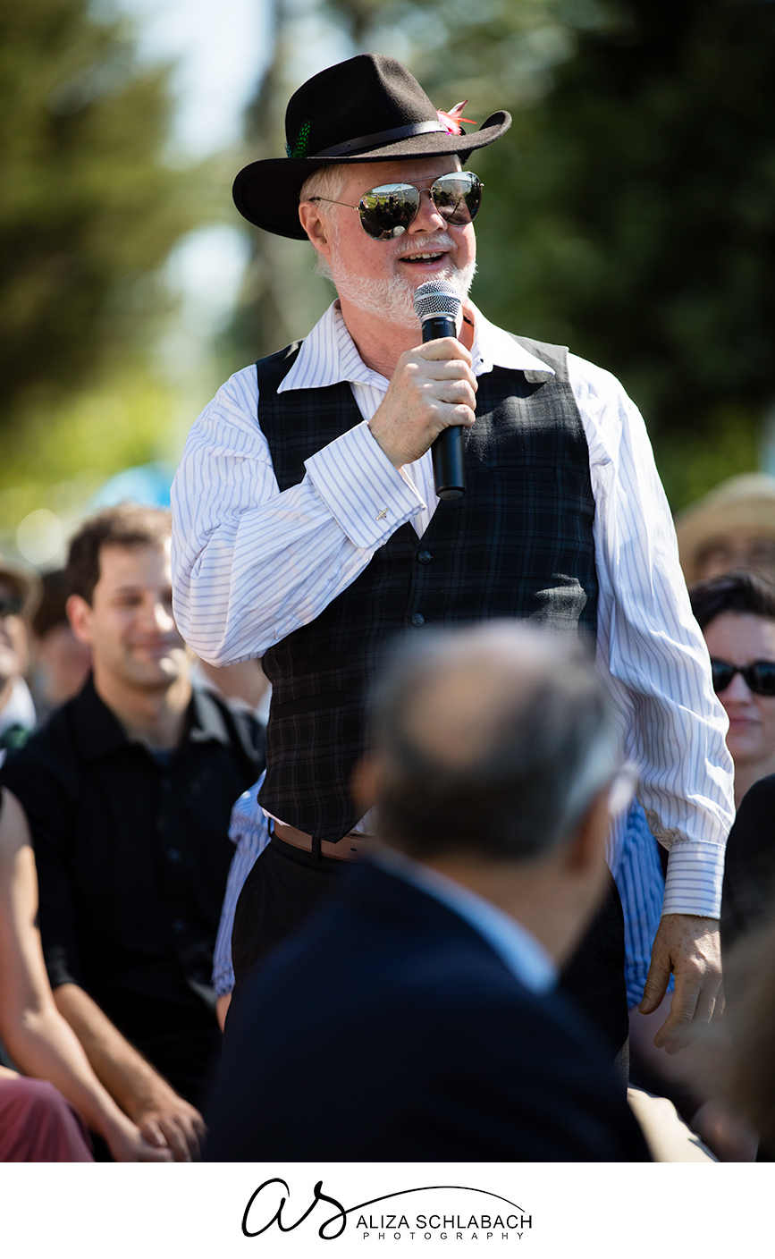Photo of a man in a had with sunglasses speaking into a microphone at a wedding ceremony