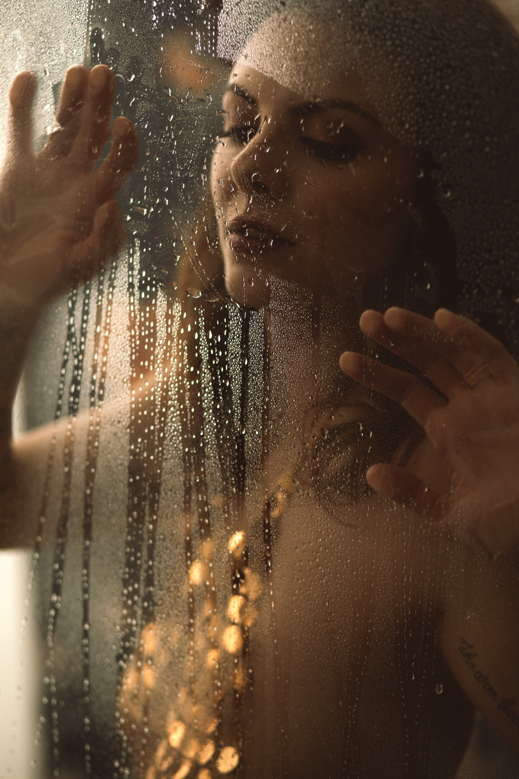 Boudoir photo of a woman in a gold sequin top behind wet shower glass