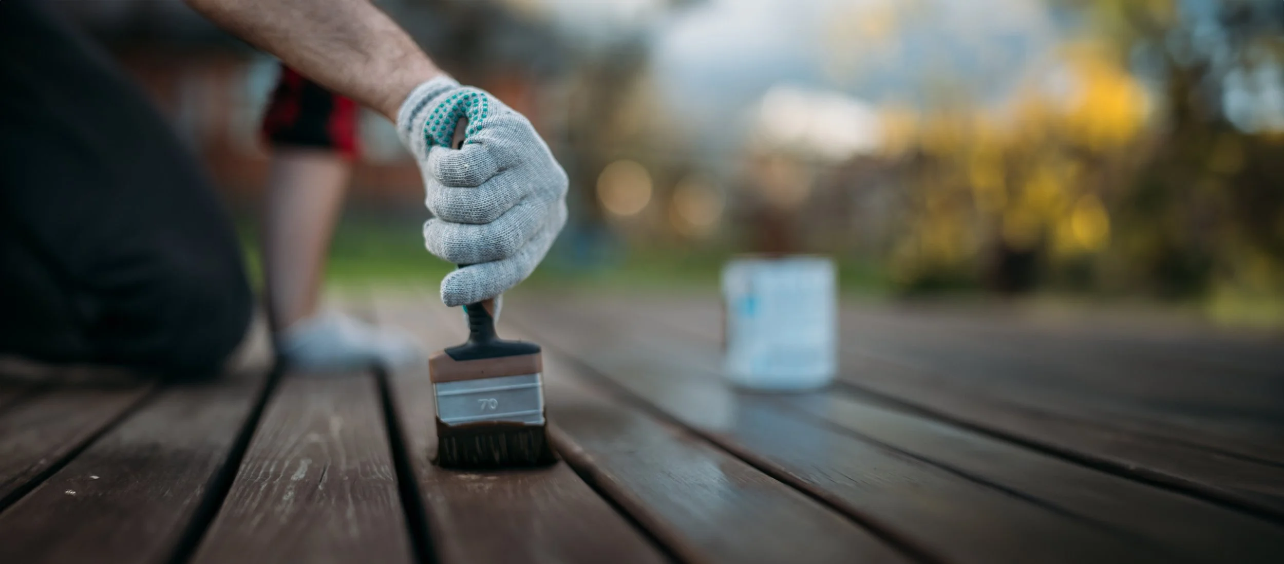 A person wearing a gray glove painting a wooden bench outdoors with a brush, with a can of paint nearby and a blurred background of trees and sky.