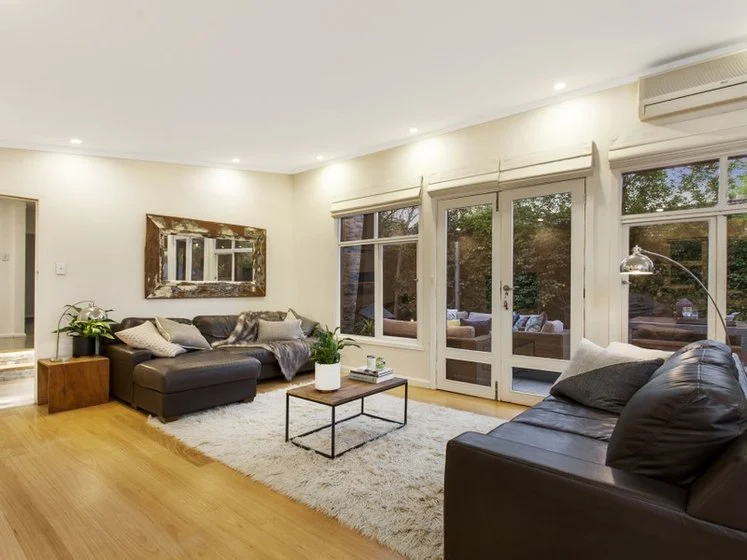 Living room with leather sofas, a coffee table, a large mirror, plants, and large windows showing trees outside.