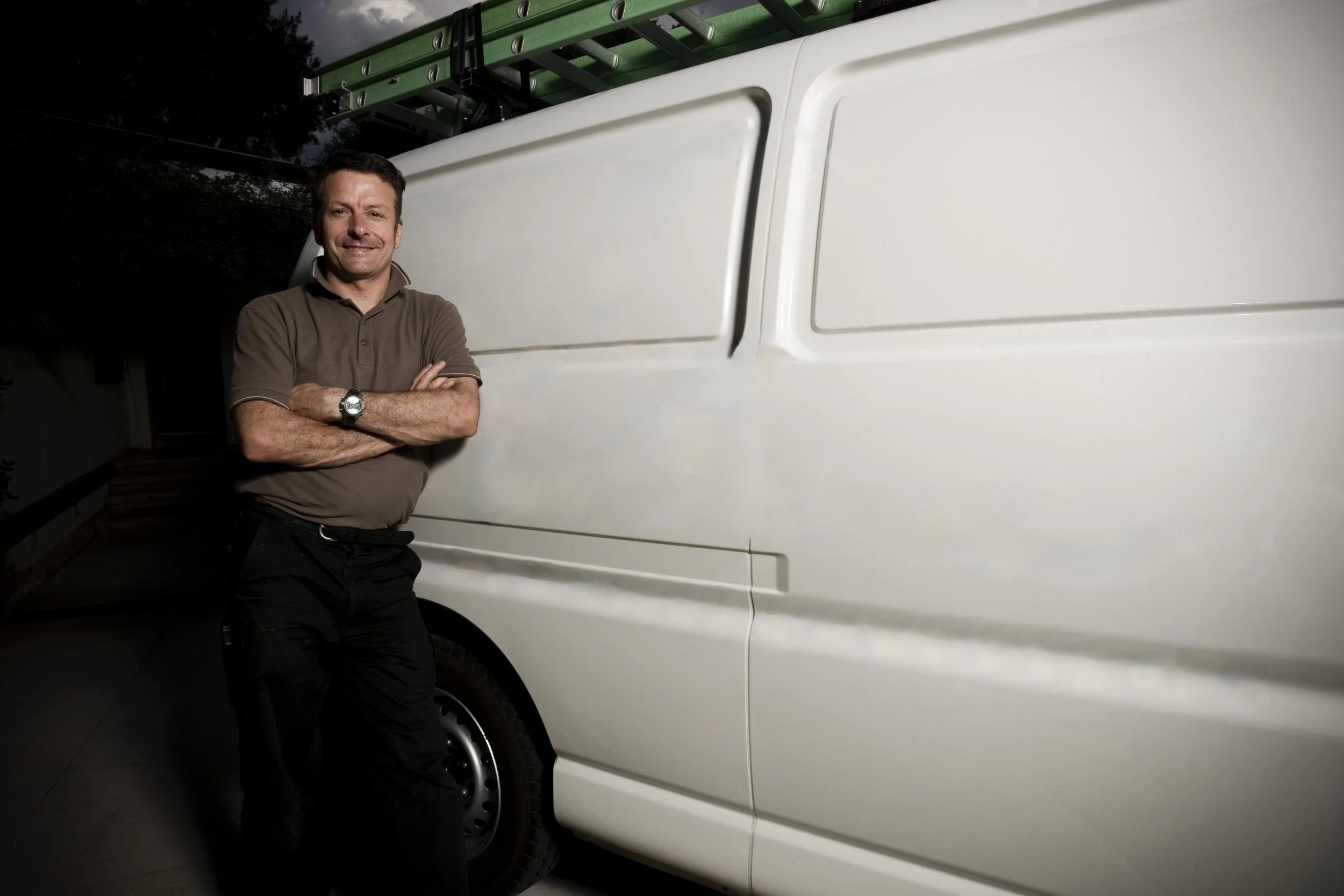 Man standing with arms crossed next to a white utility van with a ladder on top
