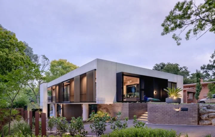 Modern two-story house with large glass windows, surrounded by greenery and trees.
