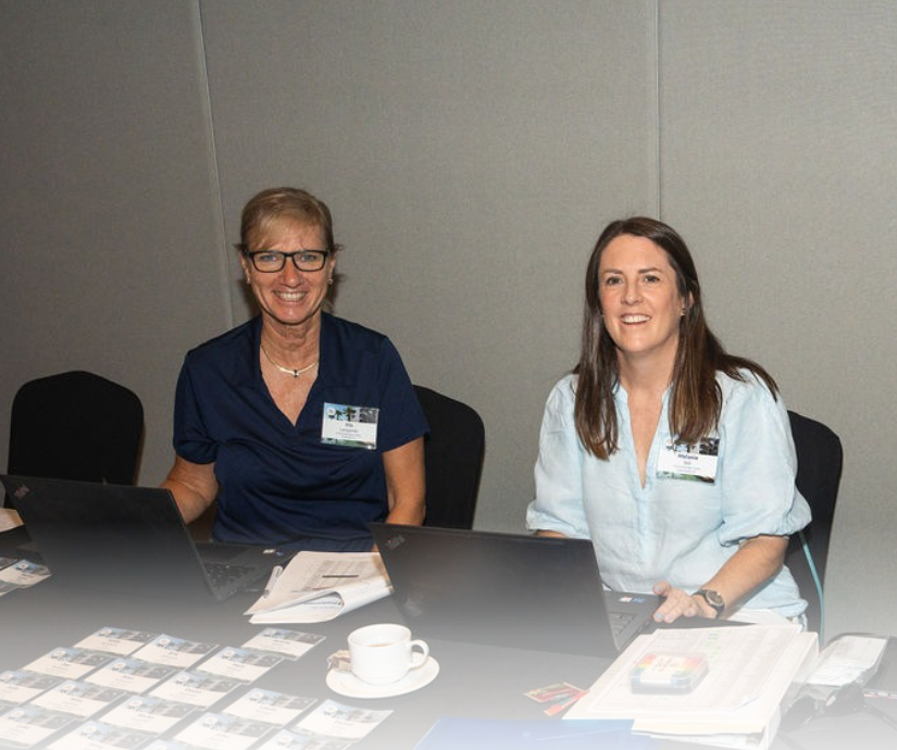 Two women sitting at a conference table with laptops, name tags, documents, a coffee cup, and conference badges.