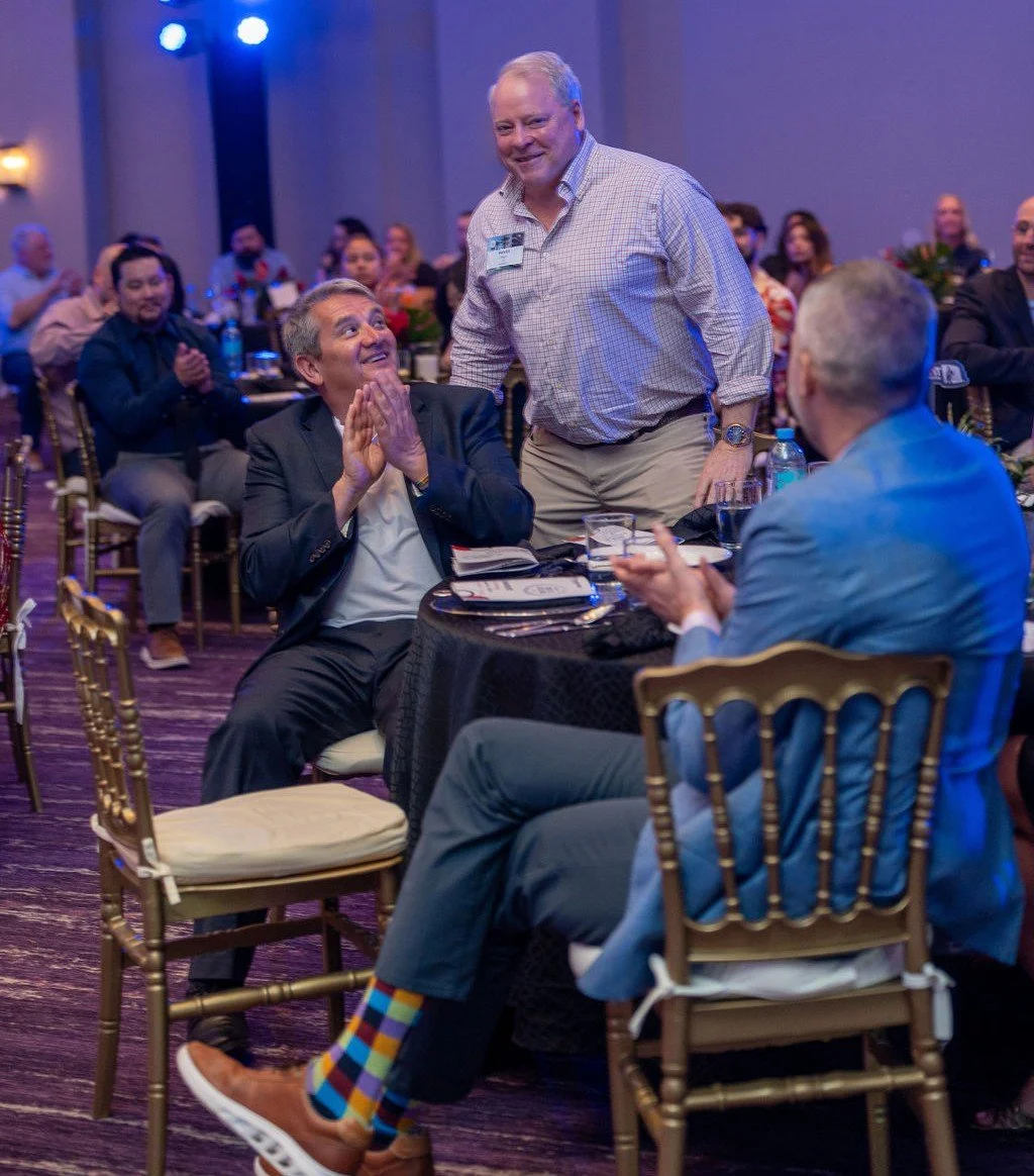 At a formal event, a man in a suit is looking up at a standing man with a smile, while the standing man is smiling back. They are seated at a table with other people clapping and smiling in the background.