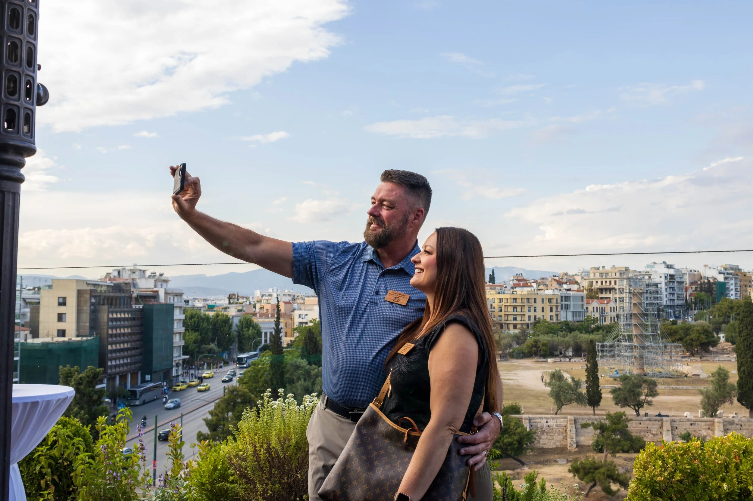 A man and woman taking a selfie on a rooftop with a cityscape in the background, smiling and posing together.