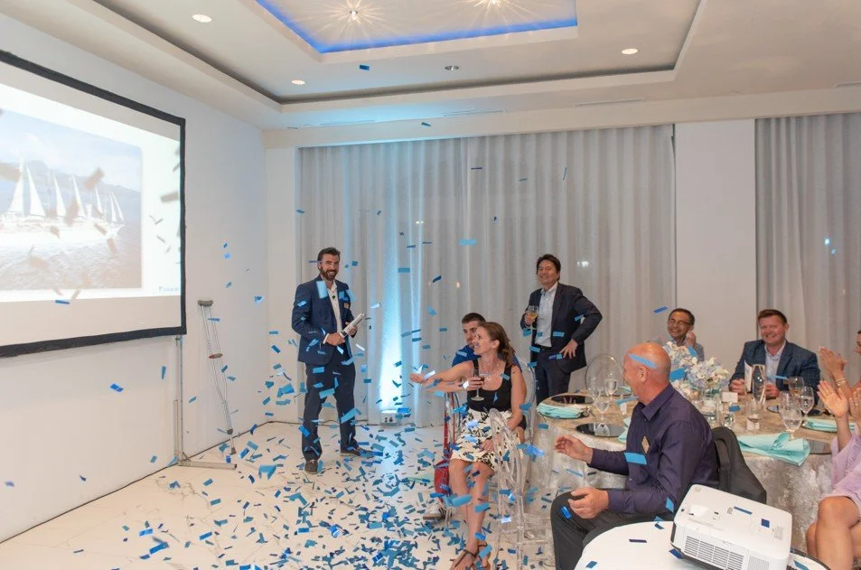 People celebrating in a banquet hall with blue confetti, some seated at round tables, others standing, one man smiling and holding a glass, and a woman sitting and reaching out