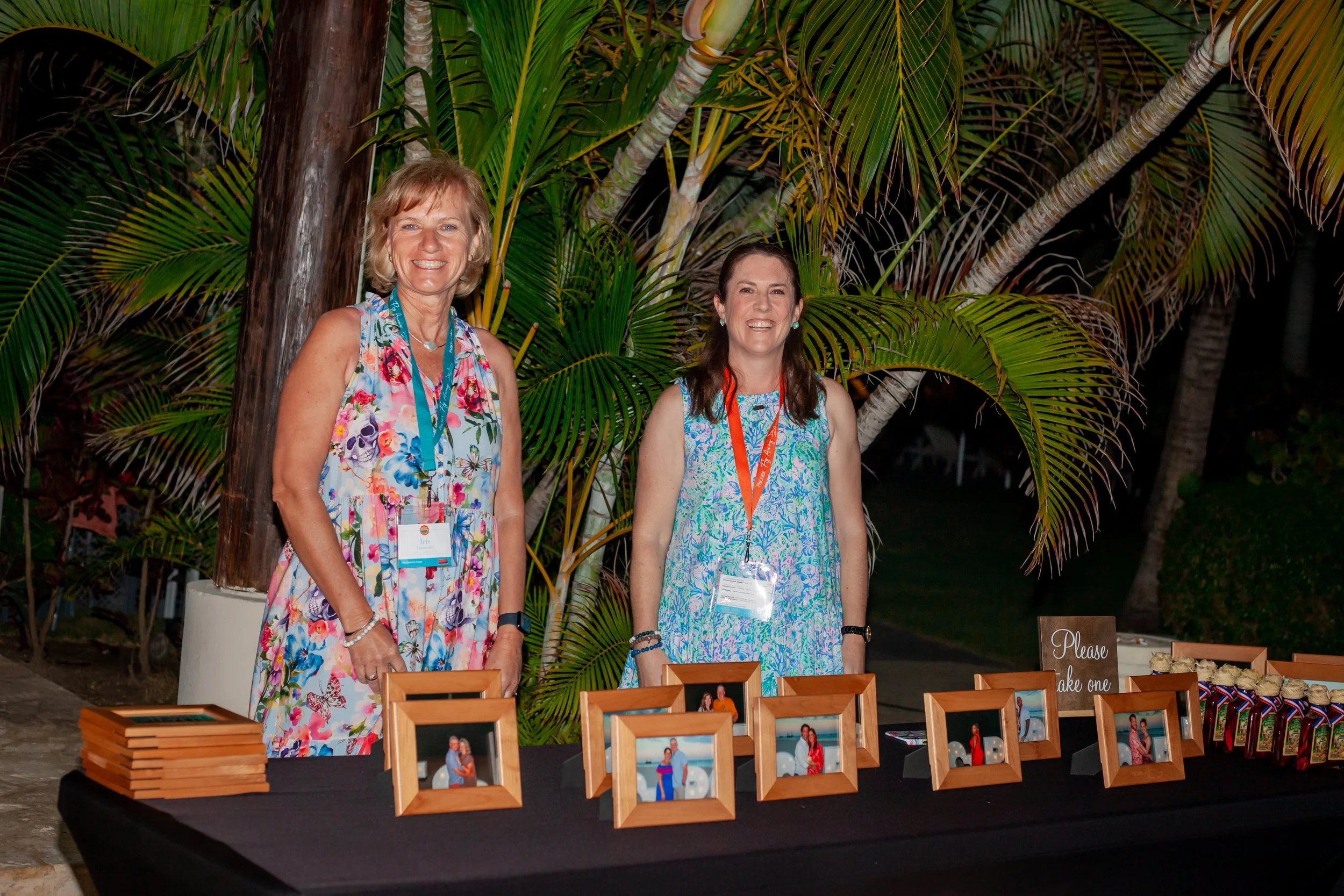 Two women in colorful dresses stand behind a table with framed photos, some baked goods, and a sign that says "Please take one" outdoors with tropical plants in the background.