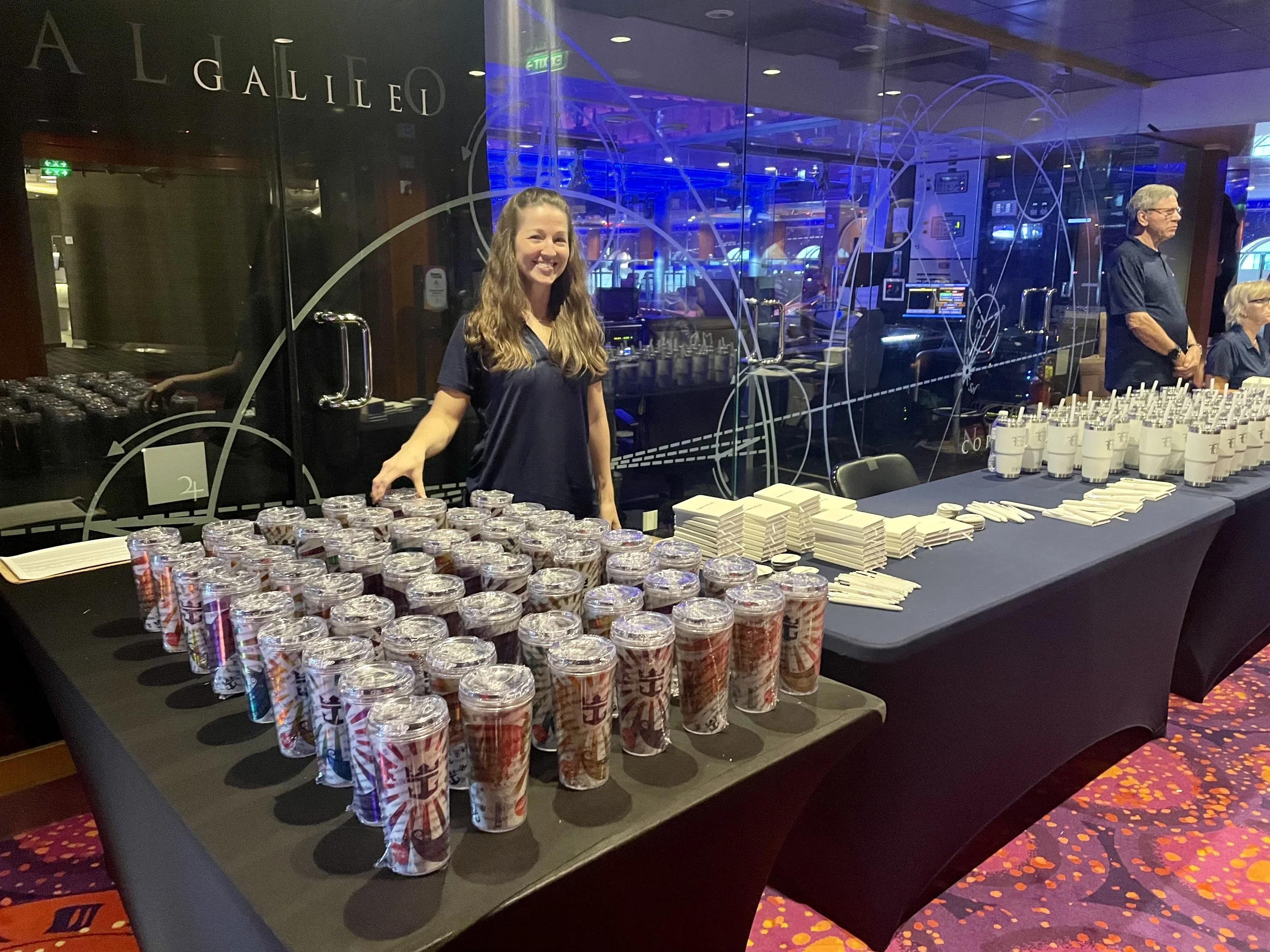 A smiling woman standing behind a table filled with themed cups at an event, with a glass wall decorated with white scientific diagrams and a view of a well-lit entertainment area in the background.