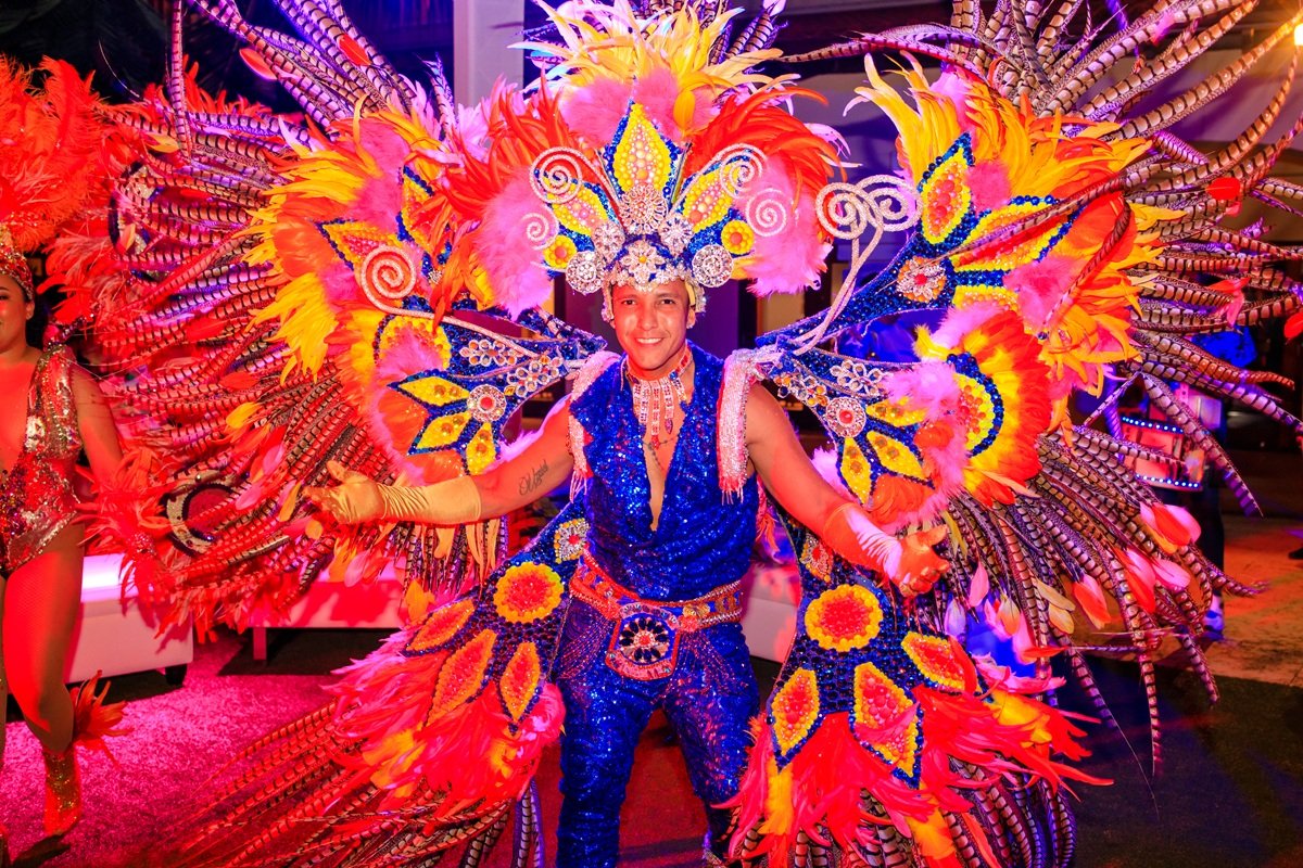 Man dressed in an elaborate, colorful carnival costume with large feathers and beadwork, smiling at the camera.