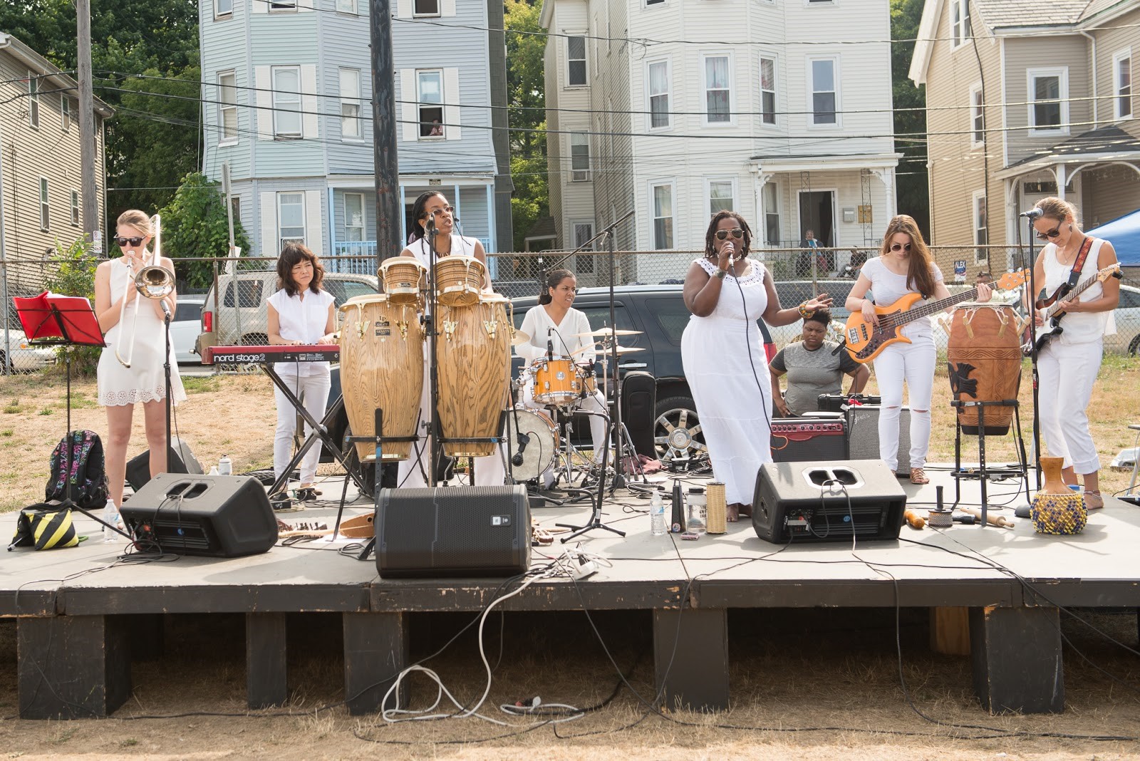 Zili Misik, a multi-ethnic all female band, lead by Kera Washington, perform at DSNI’s 28th Annual Multicultural Festival. Image shows 7 women dressed all in white singing and playing trumpet, keyboard, drum kit, bongos, guitar, and bass, on a platform at Mary Hannon Park in Dorchester. Photography by Mark Fusco.