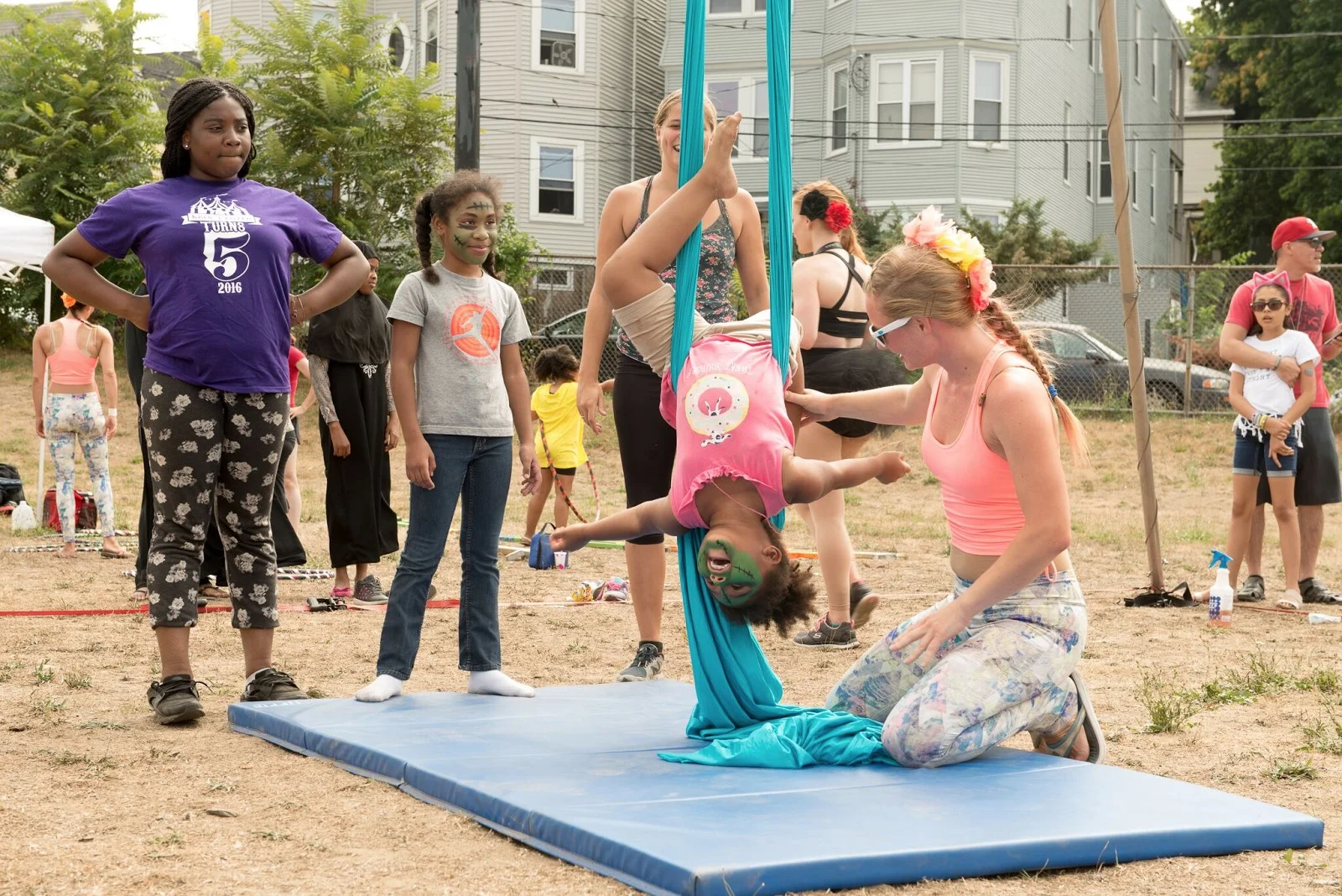 A Circus Up performer teaches a little girl at DSNI’s 28th Annual Multicultural Festival. Image shows a little girl, with her face painted, hanging upside down from blue fabric, being helped by a performer at Mary Hannon Park in Dorchester. Photography by Mark Fusco.
