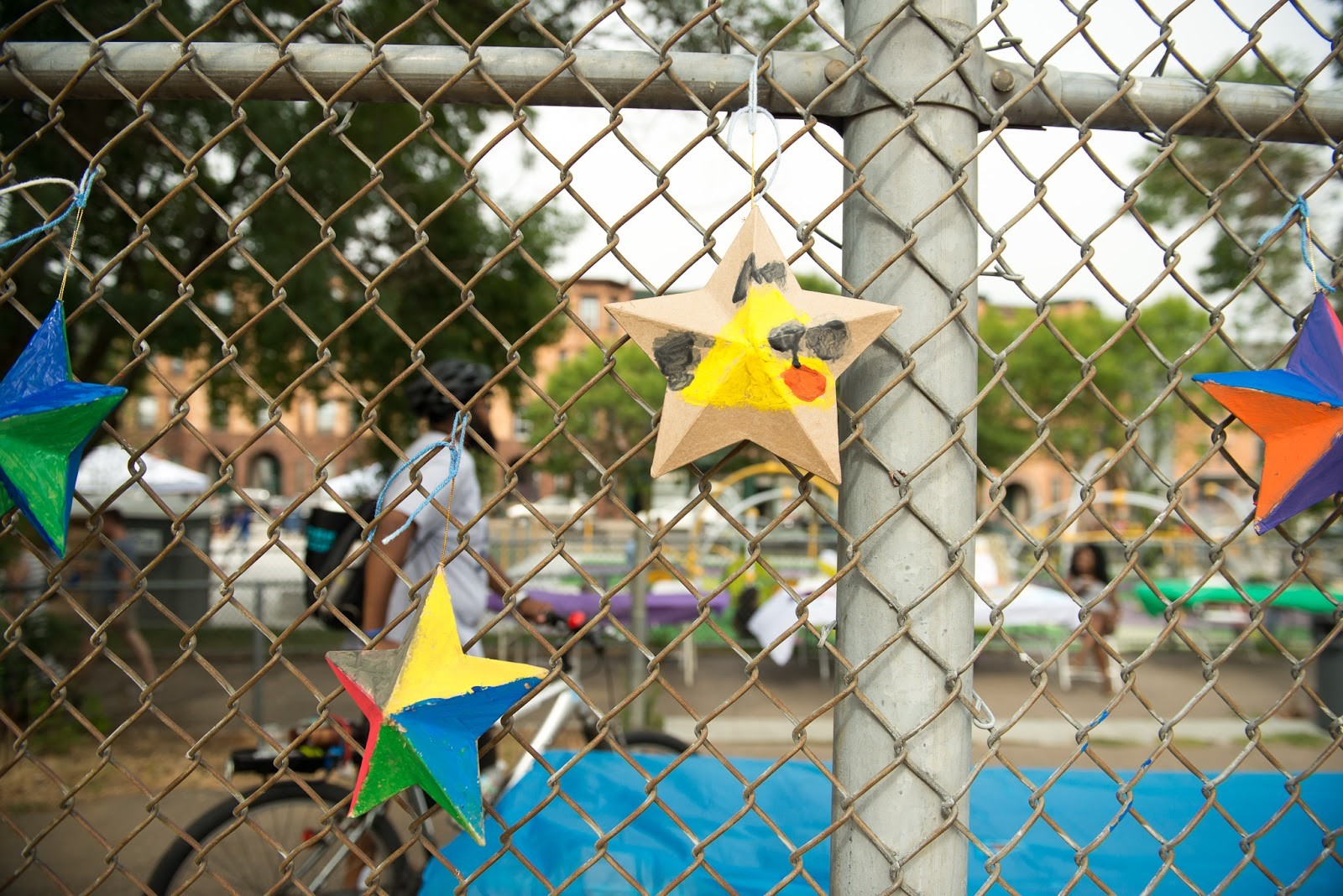 DSNI Art Wall display at 28th Annual Multicultural Festival. Image shows 4 decorated cardboard stars hanging on a chain link fence, centered is one decorated with a yellow bird. Photography by Mark Fusco.