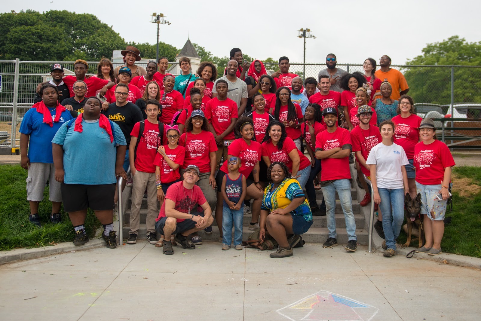 DSNI Summer Youth Organizers, Staff, and volunteers celebrate a successful 28th Annual Multicultural Festival. Image shows about 50 people, most in red T-Shirts, and a German Shepherd at Mary Hannon Park in Dorchester. Photography by Mark Fusco.