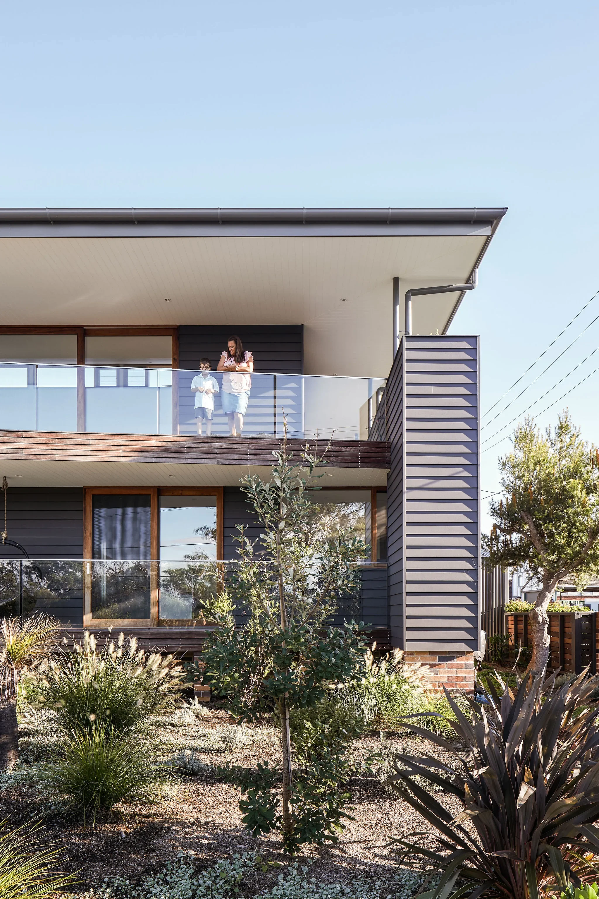 Architecturally Designed Modern Australian House with Native Gardens. Mother and son leaning on glass balustrade on balcony overlooking native Australian gardens.