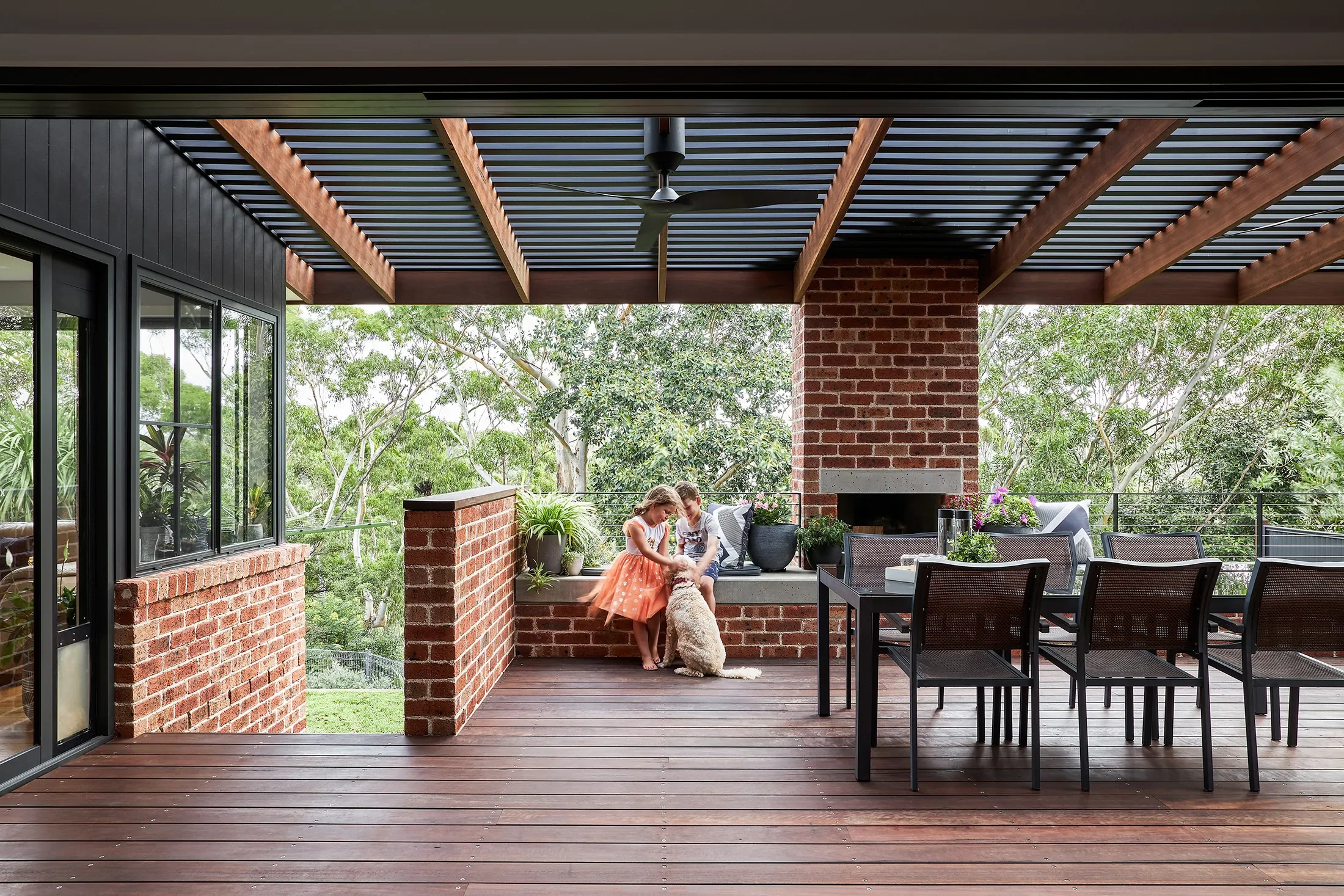 Outdoor entertaining area with children, dog, brick fireplace. Timber battens on ceiling, timber deck. 