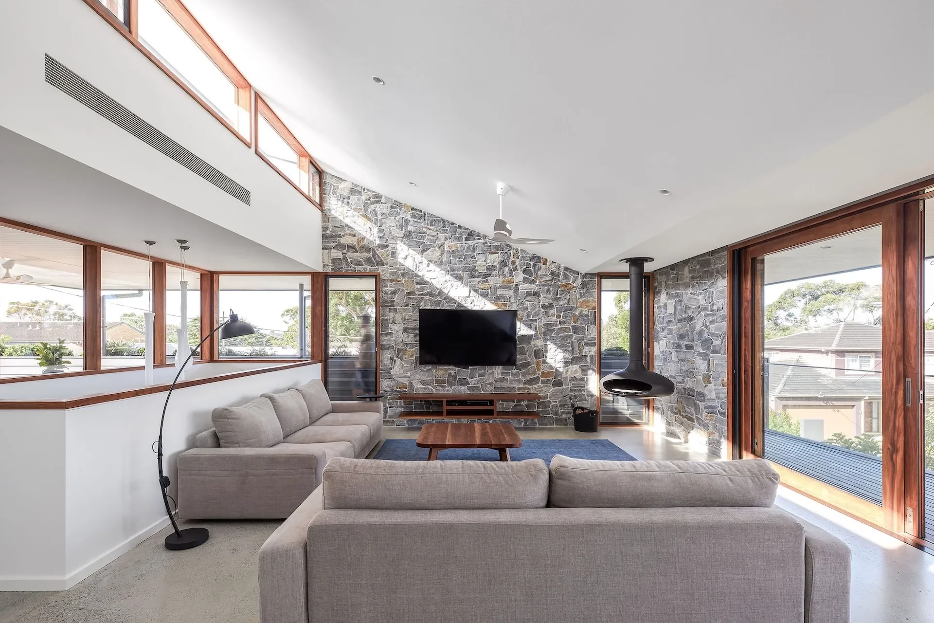 Open-plan living room with stone feature wall and large windows overlooking a leafy St Ives setting.