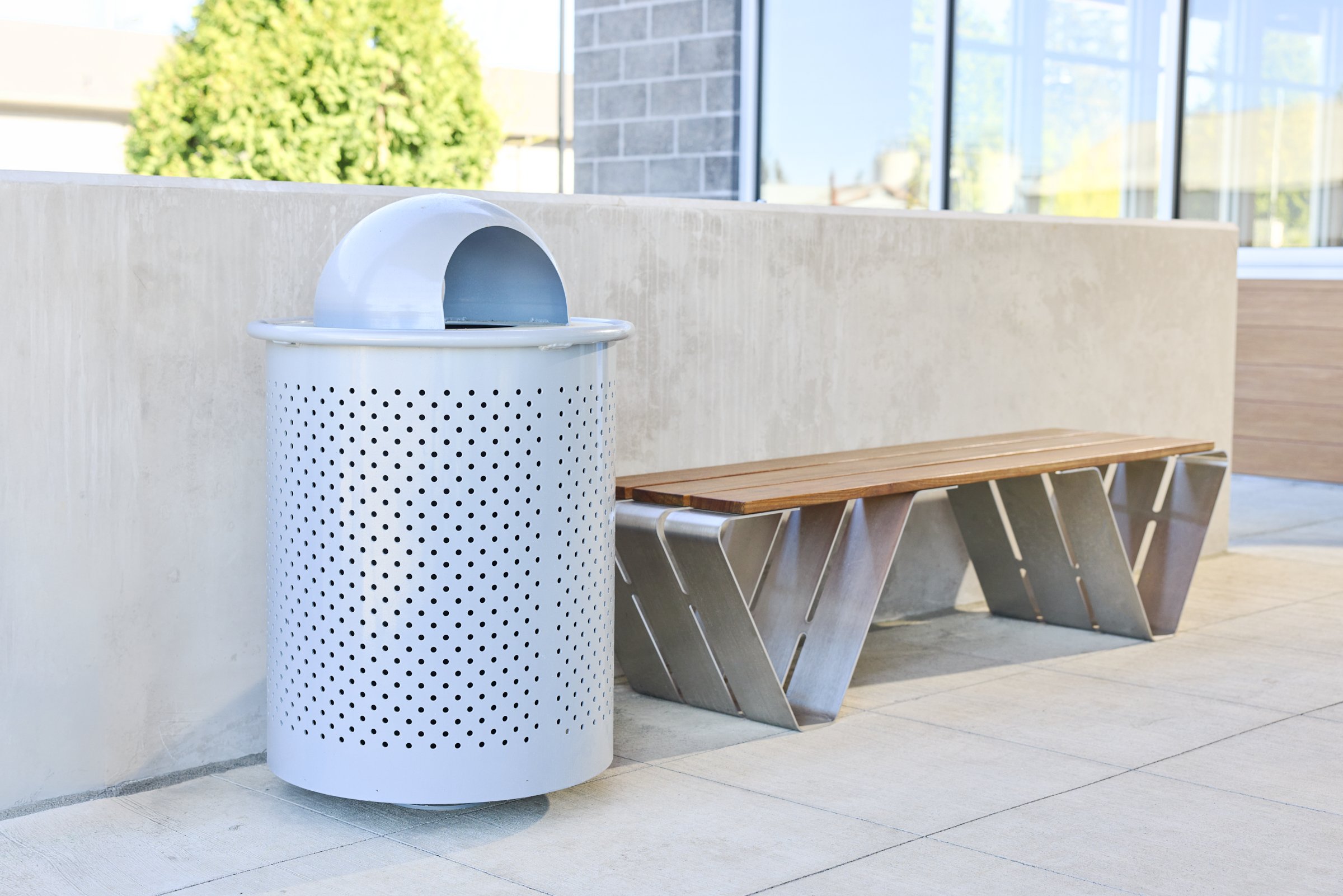 A covered Sherwood receptacle with a Douglas Bench next to it. An outdoor space with cement surrounds next to a building.