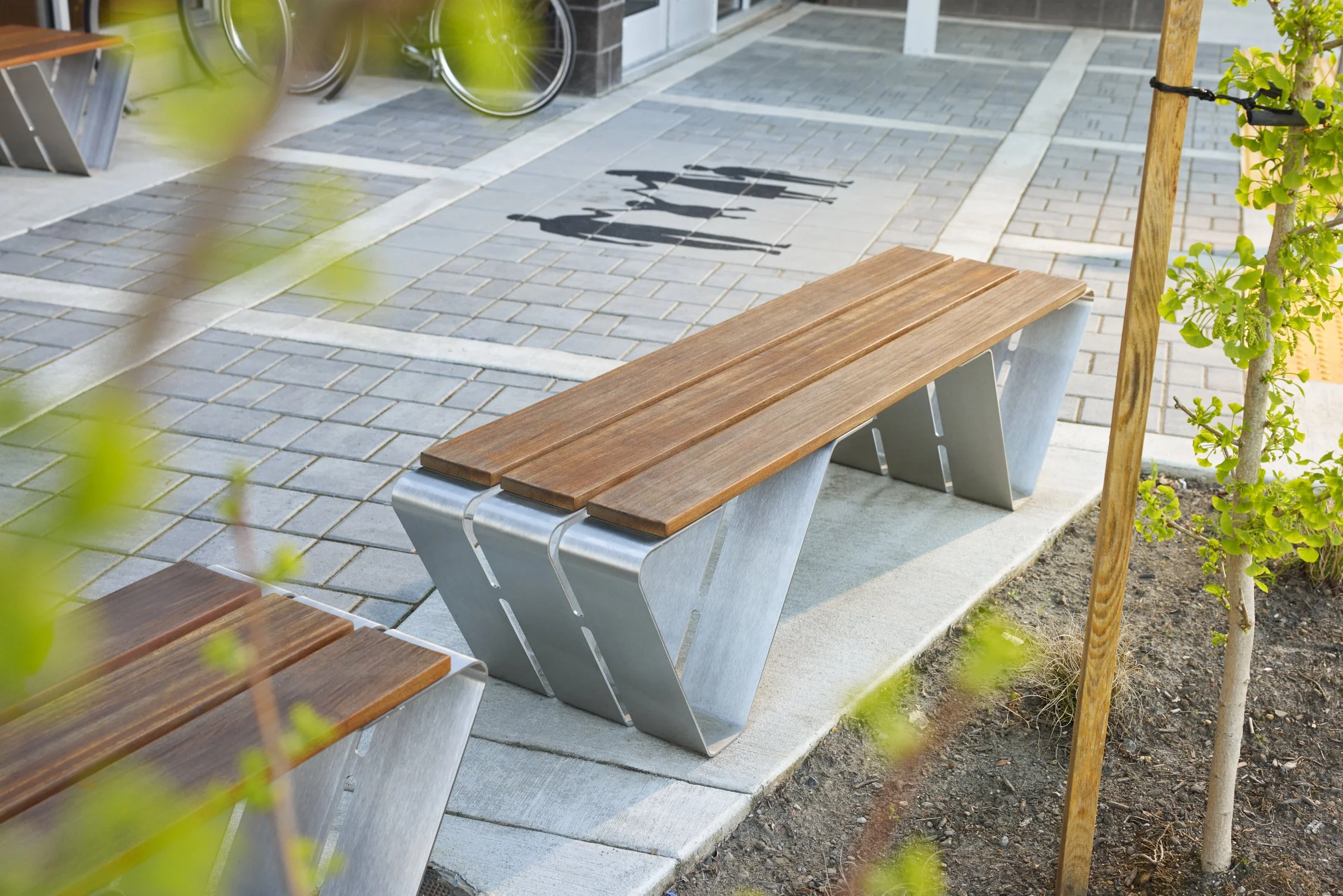 An outdoor patio in a commercial space with small trees and plantings, in the foreground are two steel base and wood-seat Douglas benches facing the building, one bench next to the building is next to a pair of Sellwood bike racks.