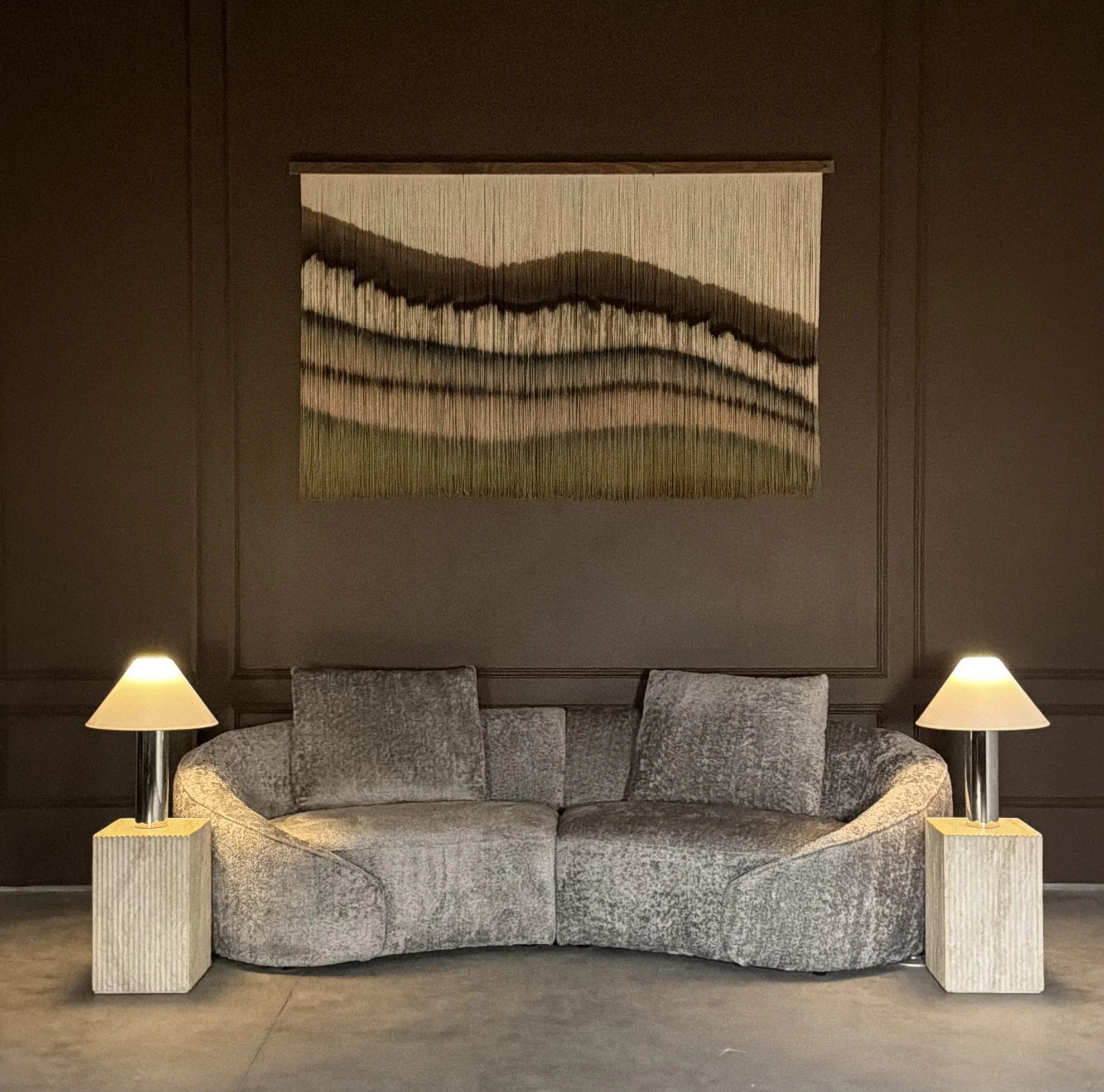 contemporary textile artwork on brown wall with gray chenille sofa and chrome side table lamps