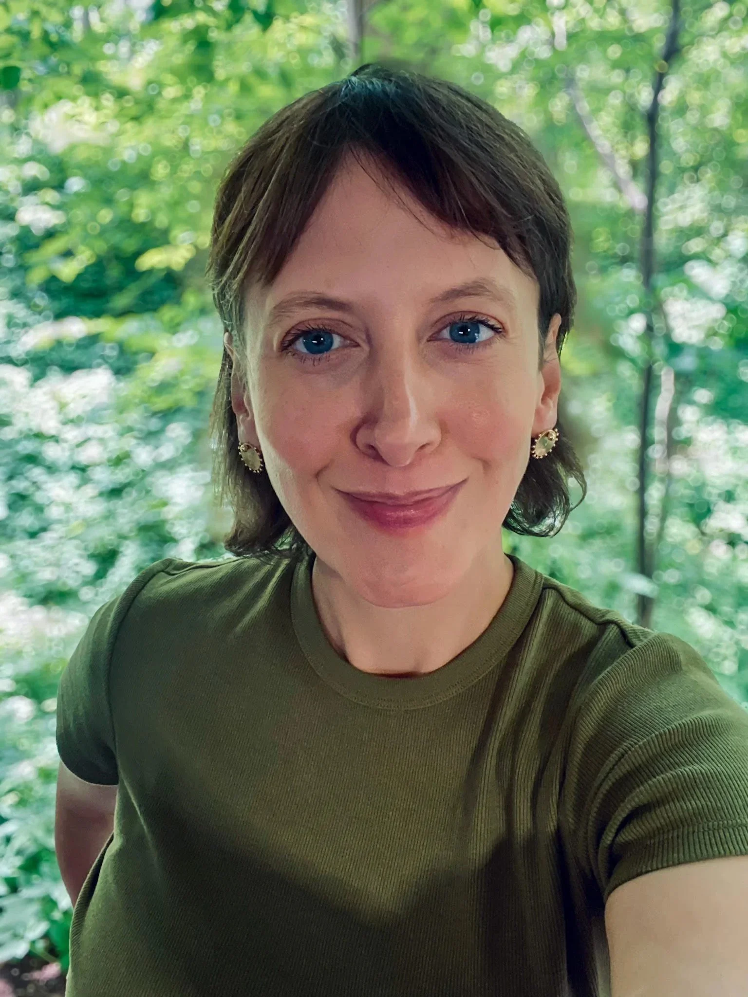 A woman with short brown hair and blue eyes smiling at the camera, wearing a green top and earrings, with a lush green forest background.