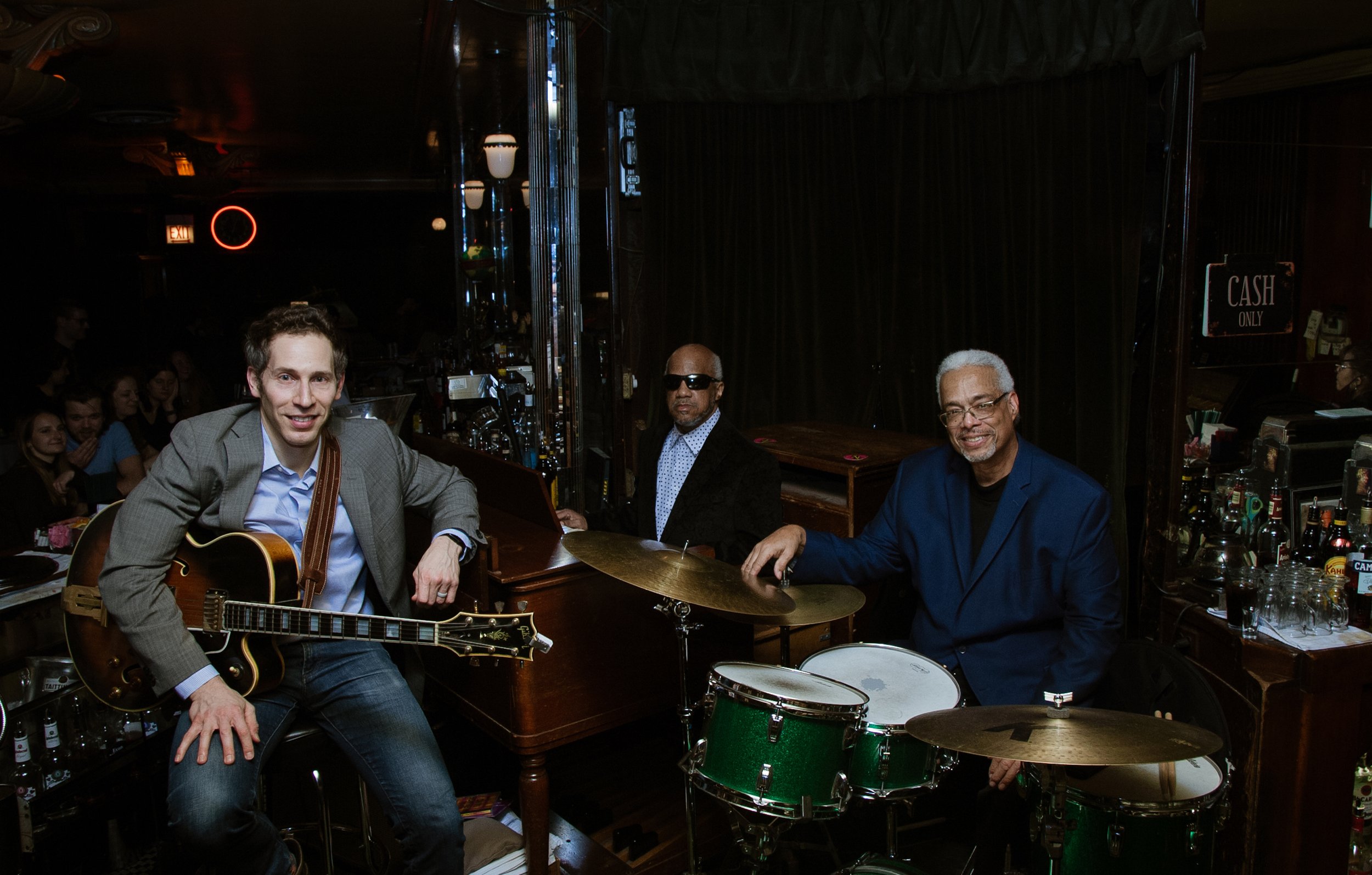Three members of the Soul Message Band posing together on stage with a bar and a crowd of people in the background.