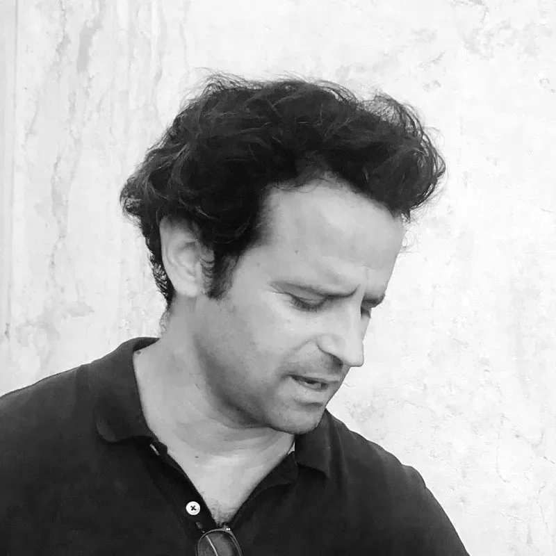 Black and white photo of a man with dark, curly hair, wearing a collared shirt, looking down with a serious expression.