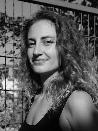Black and white photo of a young woman with curly hair smiling outdoors in front of a fence.