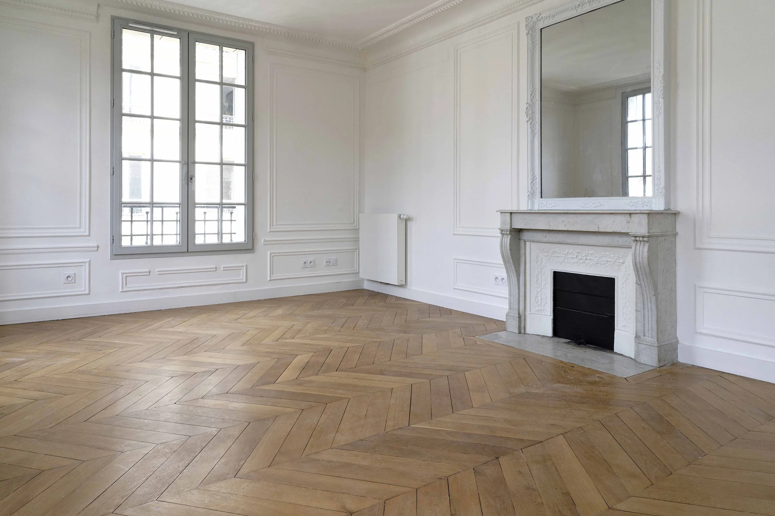 Empty elegant living room with white walls, large windows, wooden herringbone floor, decorative molding, and a white fireplace with a mirror above it.