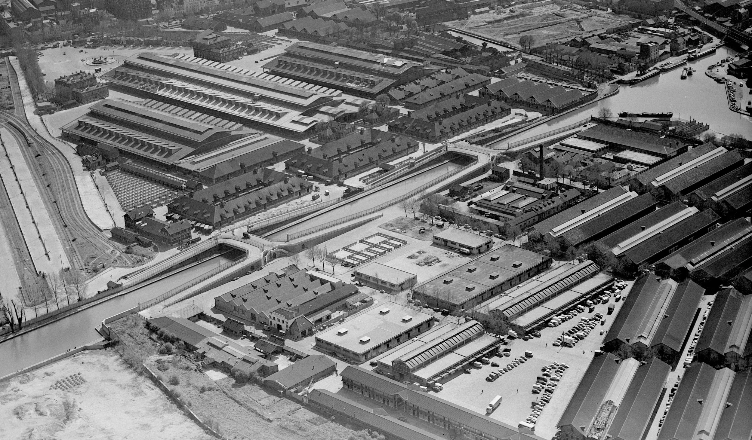 Aerial view of an urban area with industrial buildings, warehouses, houses, and a body of water with boats docked along its edge. Snow covers the ground and rooftops, indicating winter.