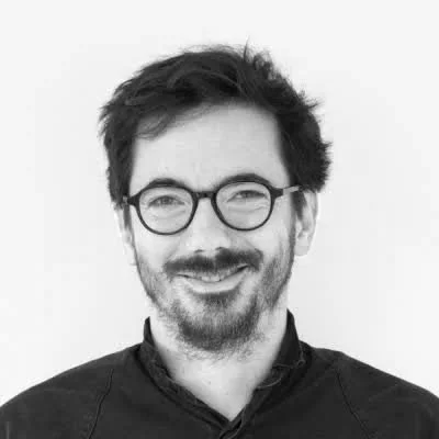 Black-and-white photo of a man with glasses, a beard, and messy hair, smiling against a plain white background.