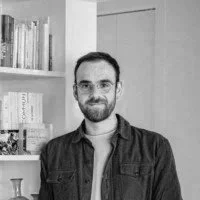 A man with glasses and a beard smiling, standing in front of a bookshelf in a room.