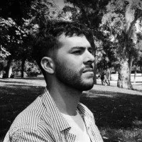 A young man with a beard and short hair, wearing a striped shirt, looking to the side outdoors in a park with trees in the background, in black and white.