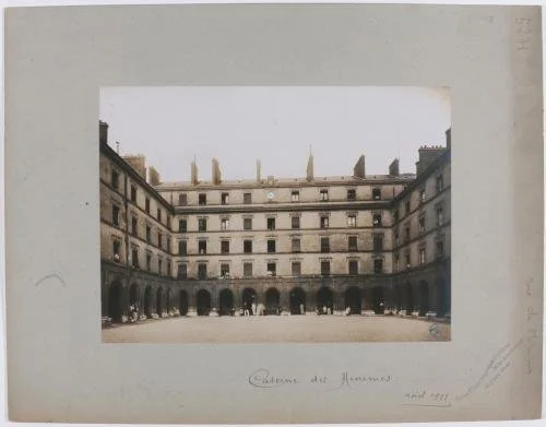 Empty courtyard surrounded by four-story historic European building with arched ground level, ornate windows, and rooftop chimneys.