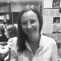 Smiling woman with glasses and long dark hair, wearing a white shirt, in a room with bookshelves and people in the background.