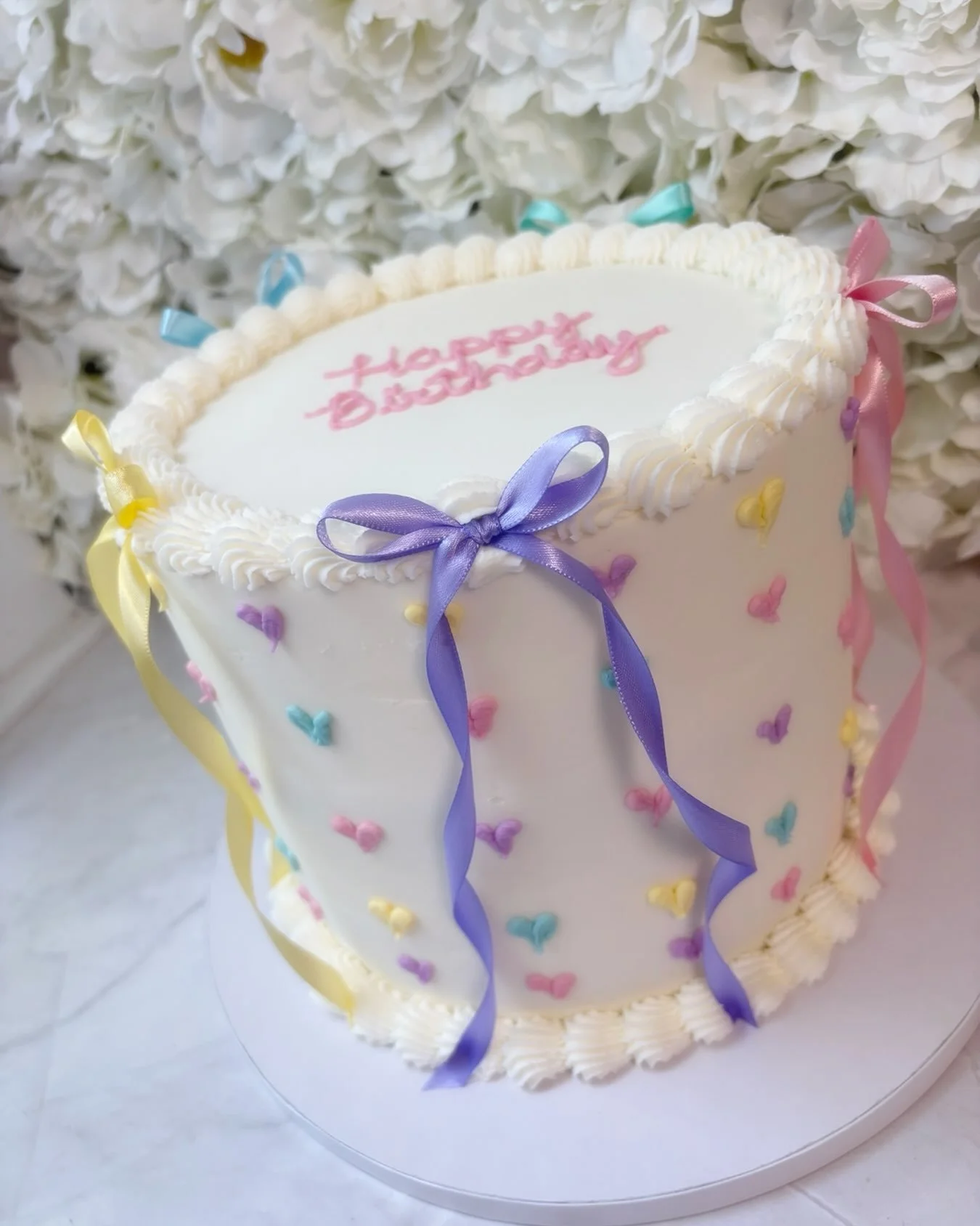 Pastel perfection for Evelyn&rsquo;s 2nd birthday
🎀💜💛🩷🩵

A sweet vintage-inspired birthday cake featuring delicate buttercream hearts, classic piped borders, and colorful satin bows for the prettiest finishing touch. Simple, playful, and made to