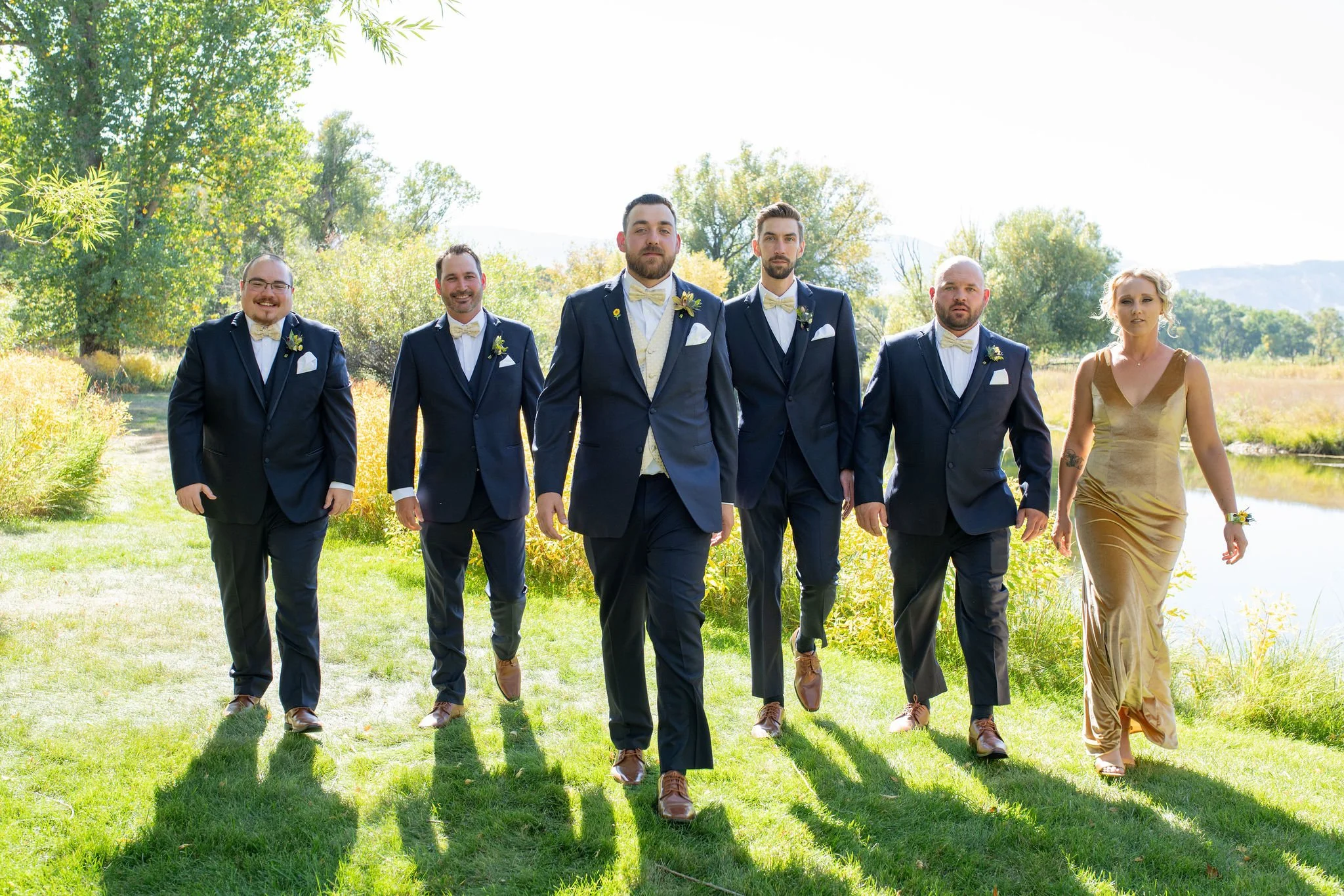 A groom walks with his attendants with the men in navy suits and his grooms-woman in a gold velvet dress for a September wedding in Wyoming