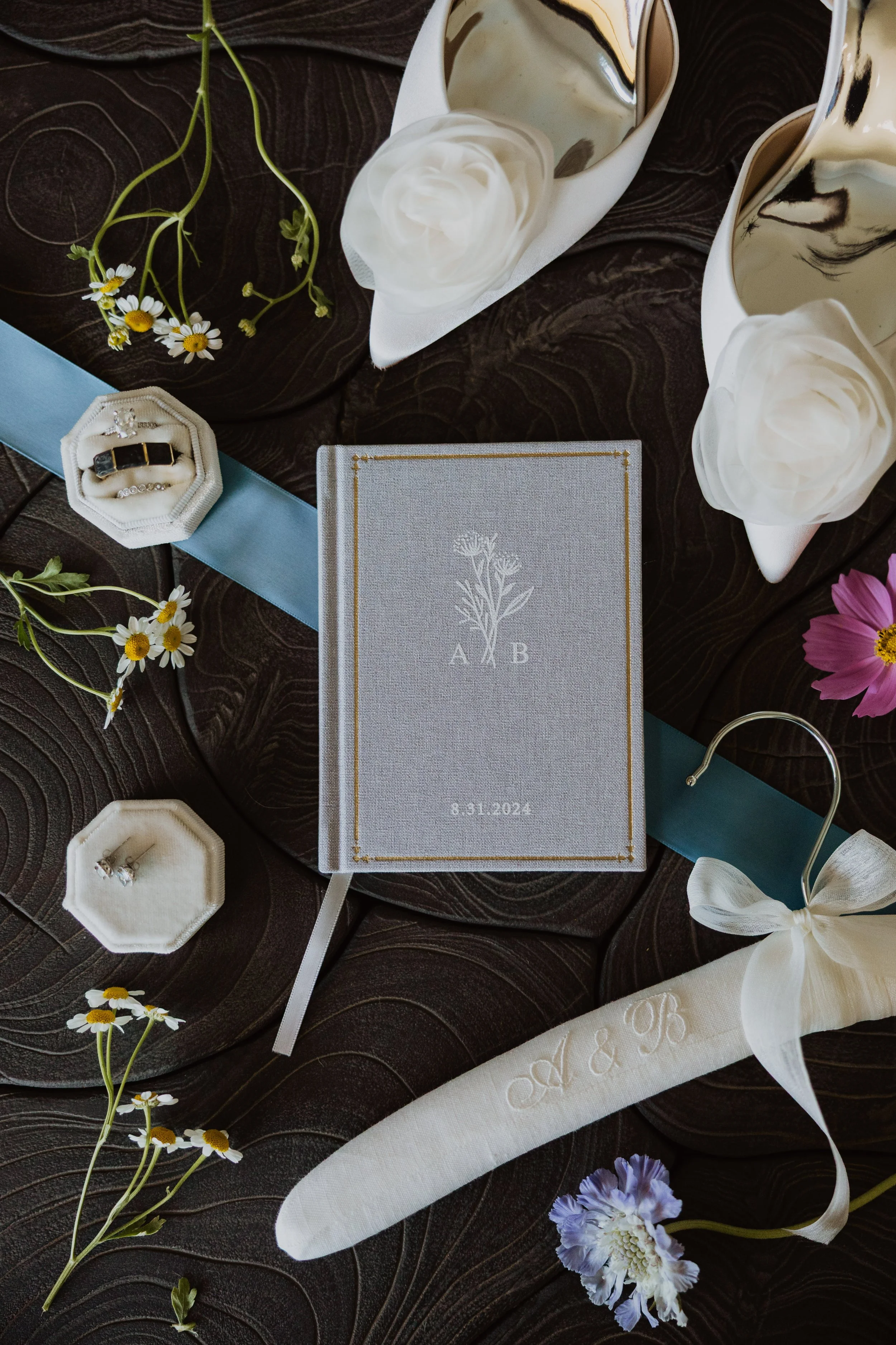 Wedding details included bridal shoes with white rosette, embroidered hair clip, wedding rings, linen vow book and diamond earrings with florals