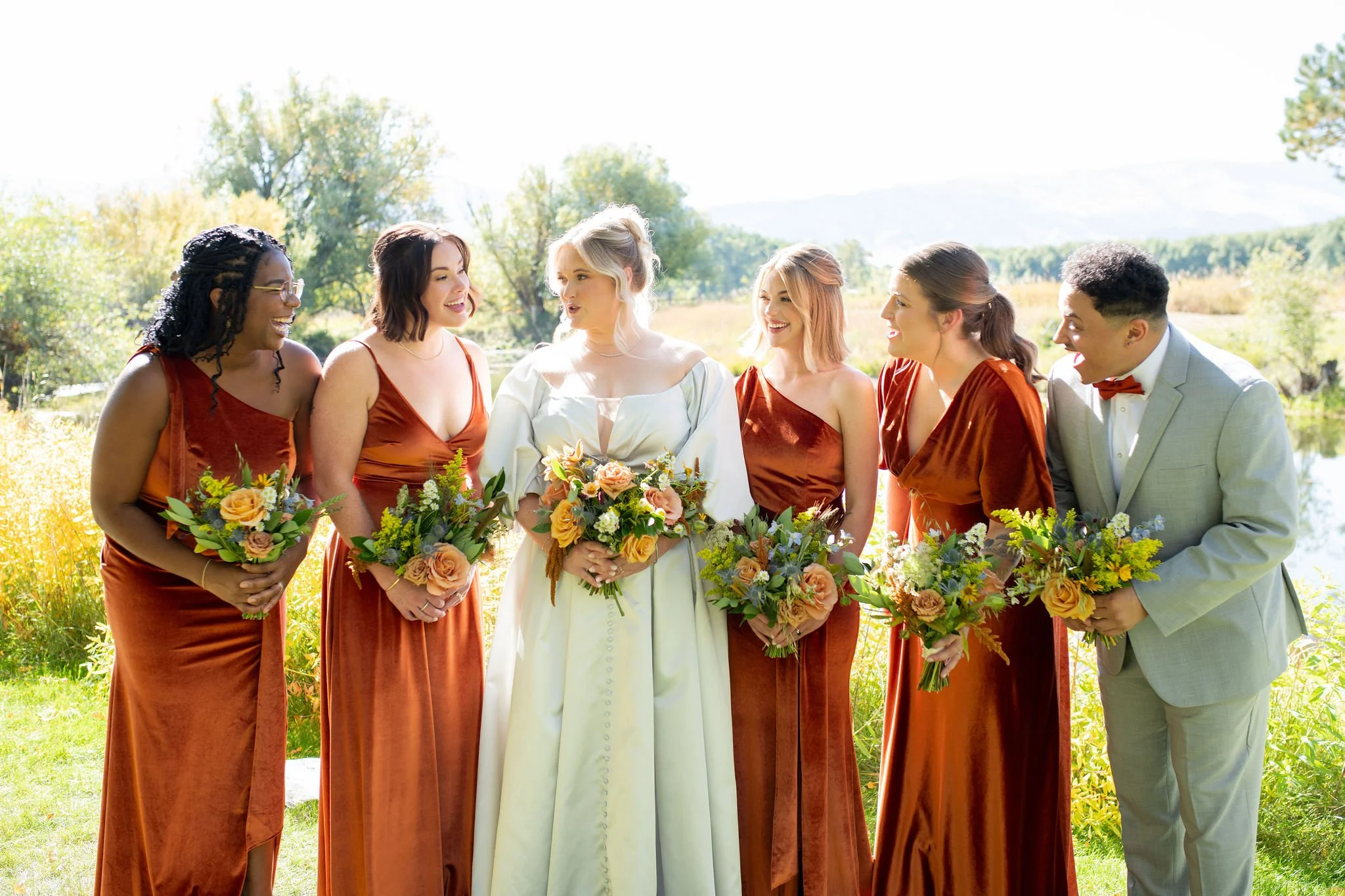 A bride stands with her mixed bridal party with the bridesmaids wearing varied styles in Burant Orange and her male attendant in a light grey suit for this September Wyoming Wedding with flowers by Whirly Girl Flowers, a Sheridan, WY wedding florist.
