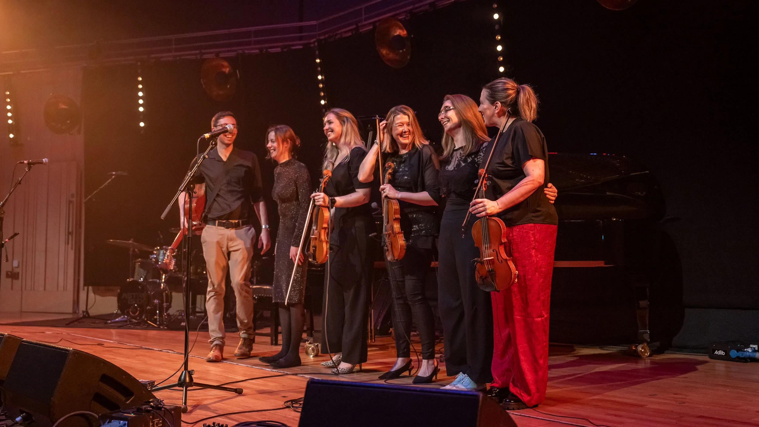 Group of seven musicians standing on stage after a performance, holding violins and smiling. They are dressed in semi-formal attire, with a grand piano in the background and stage lights overhead.