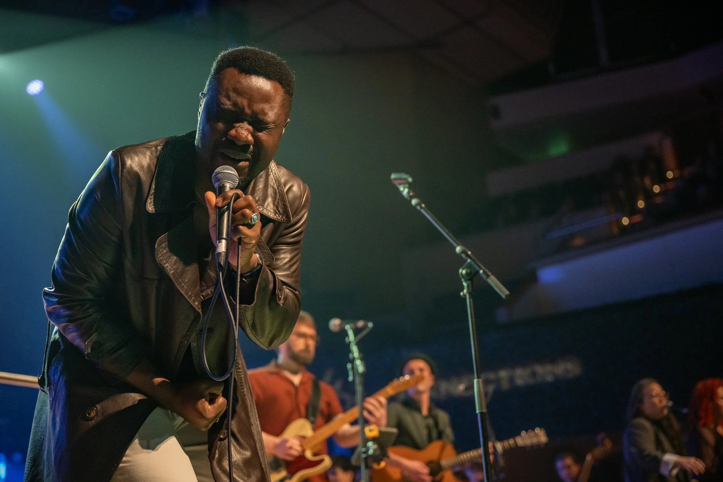 A man passionately singing into a microphone during a live music performance, with a band playing instruments in the background on stage.