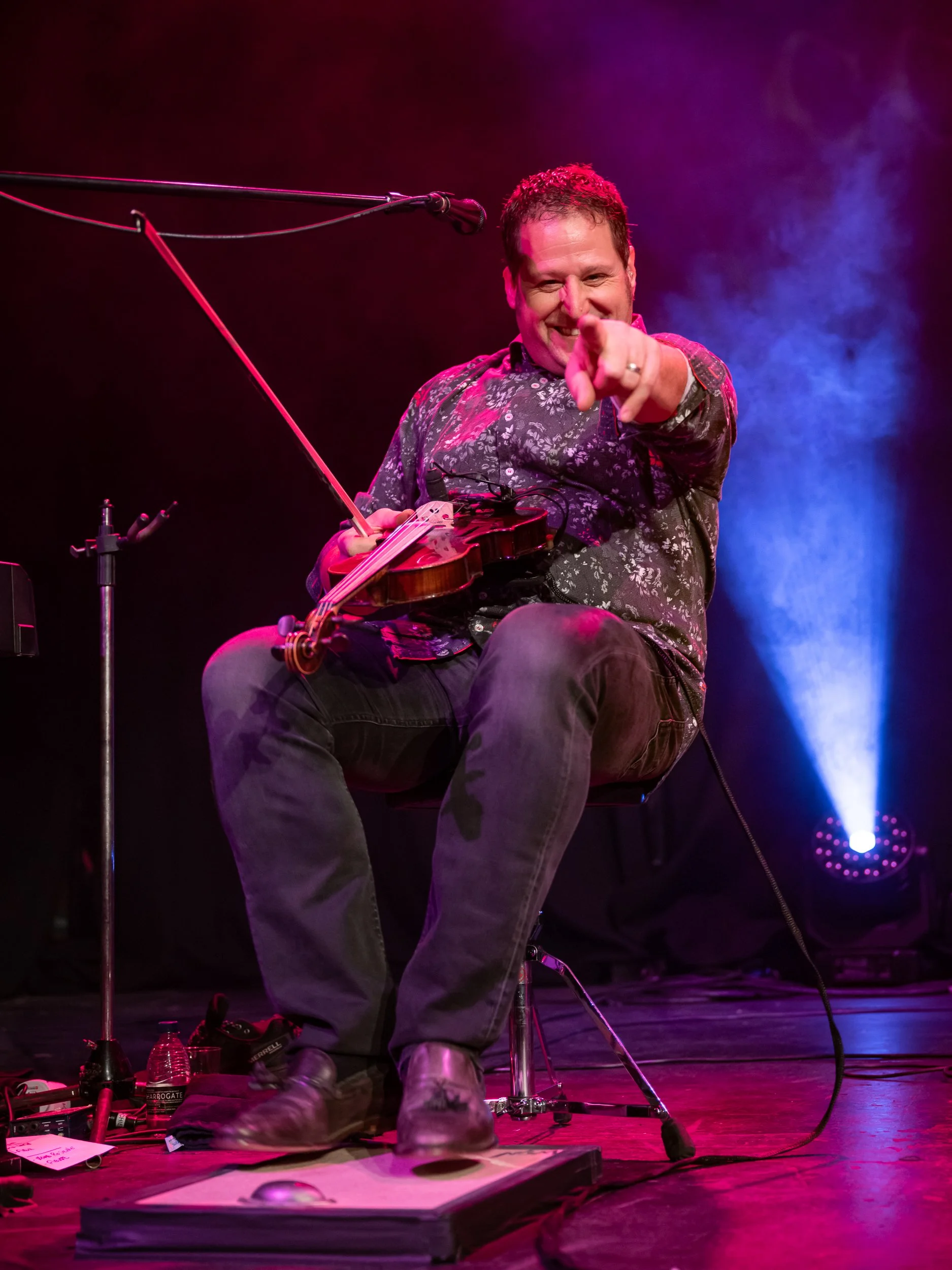 A man playing a violin on stage, pointing towards the camera, with colorful stage lighting in the background.