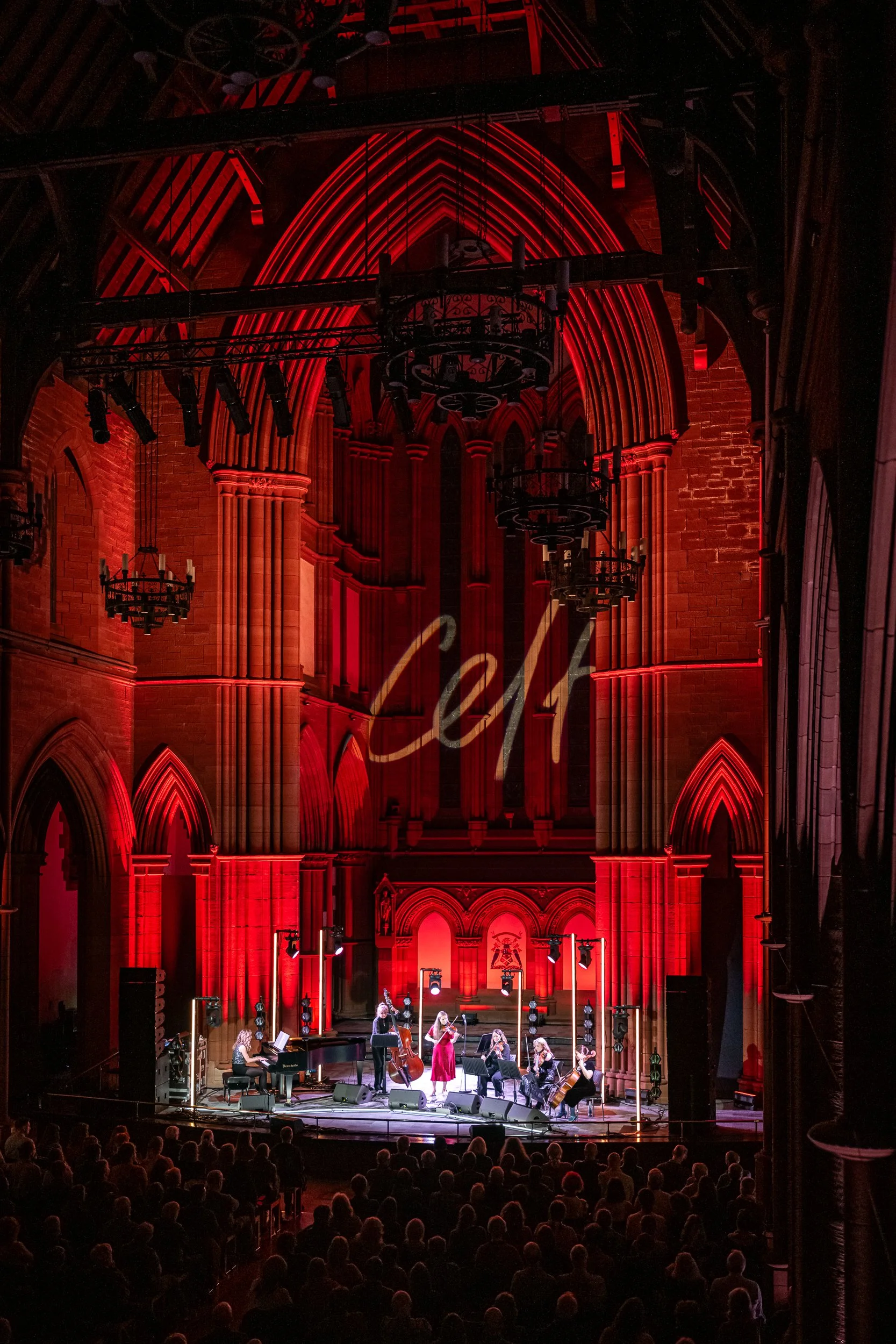 A musical performance taking place inside a large, gothic-style cathedral with red lighting. Musicians are playing instruments on stage, including a piano, cello, and violin, while an audience watches.