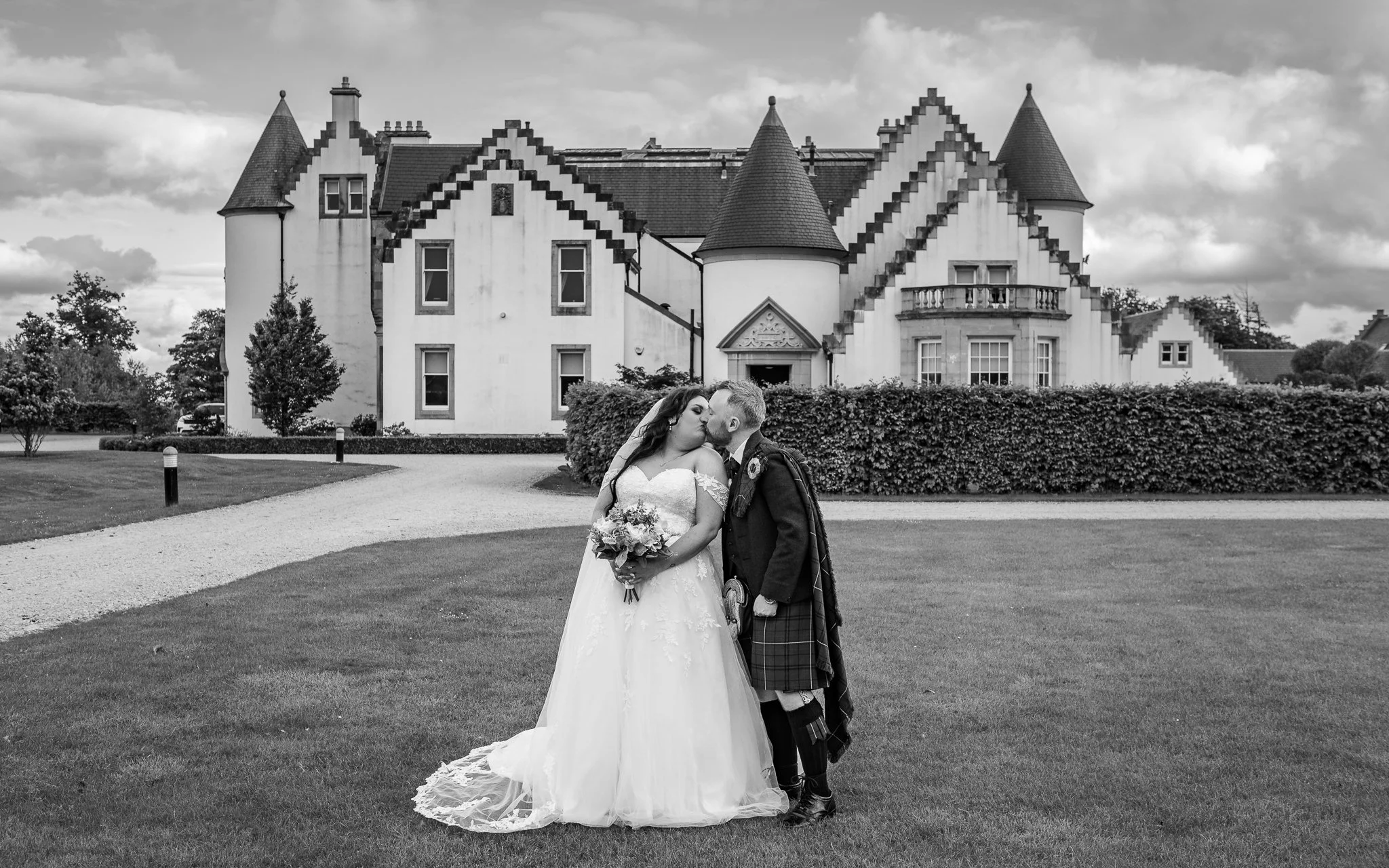 Black and white photo of a bride and groom kissing in front of a large castle-style house with turrets, surrounded by a lawn and trees.