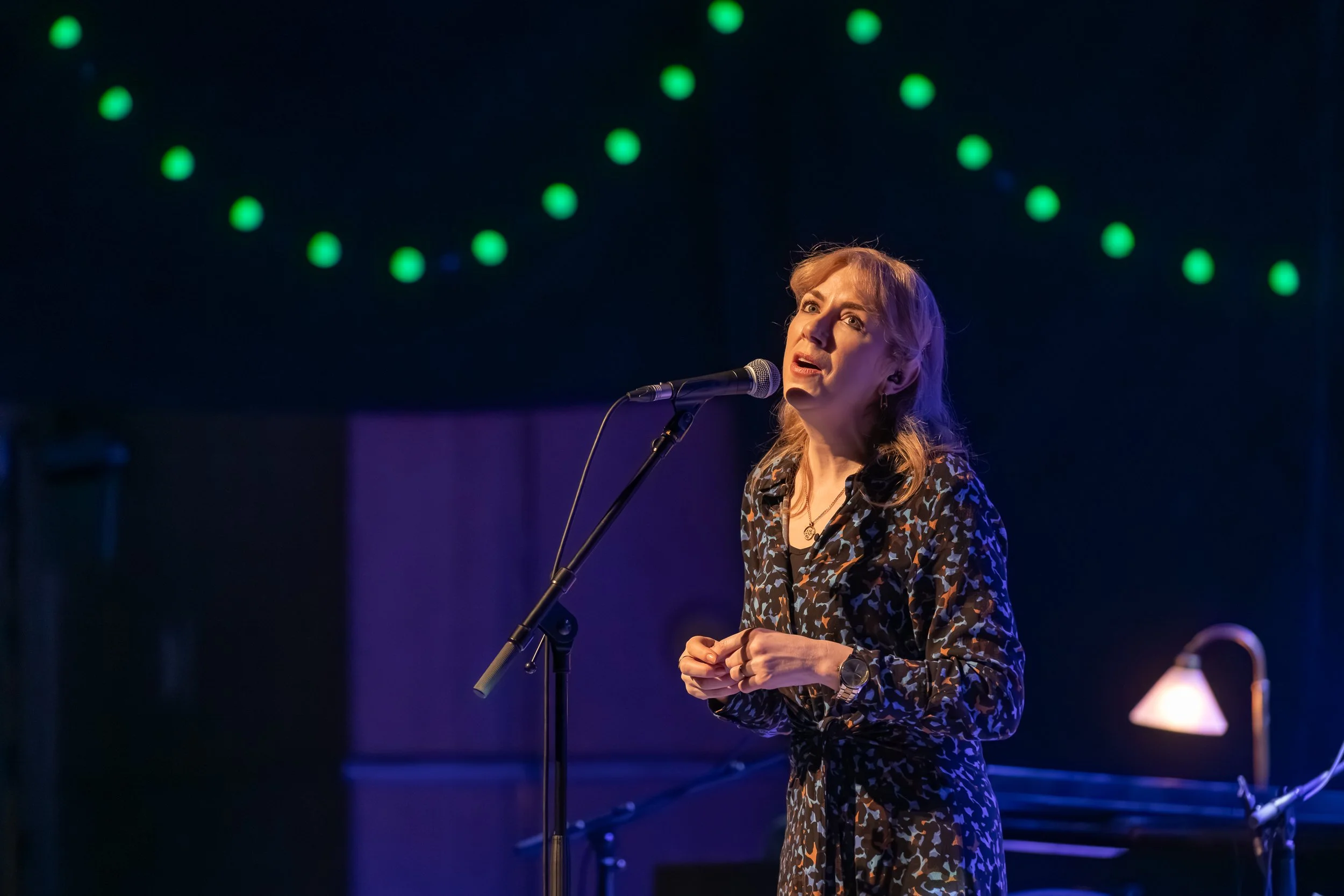 A woman performing on stage with a microphone, wearing a patterned dress, with green string lights hanging overhead and a piano in the background.