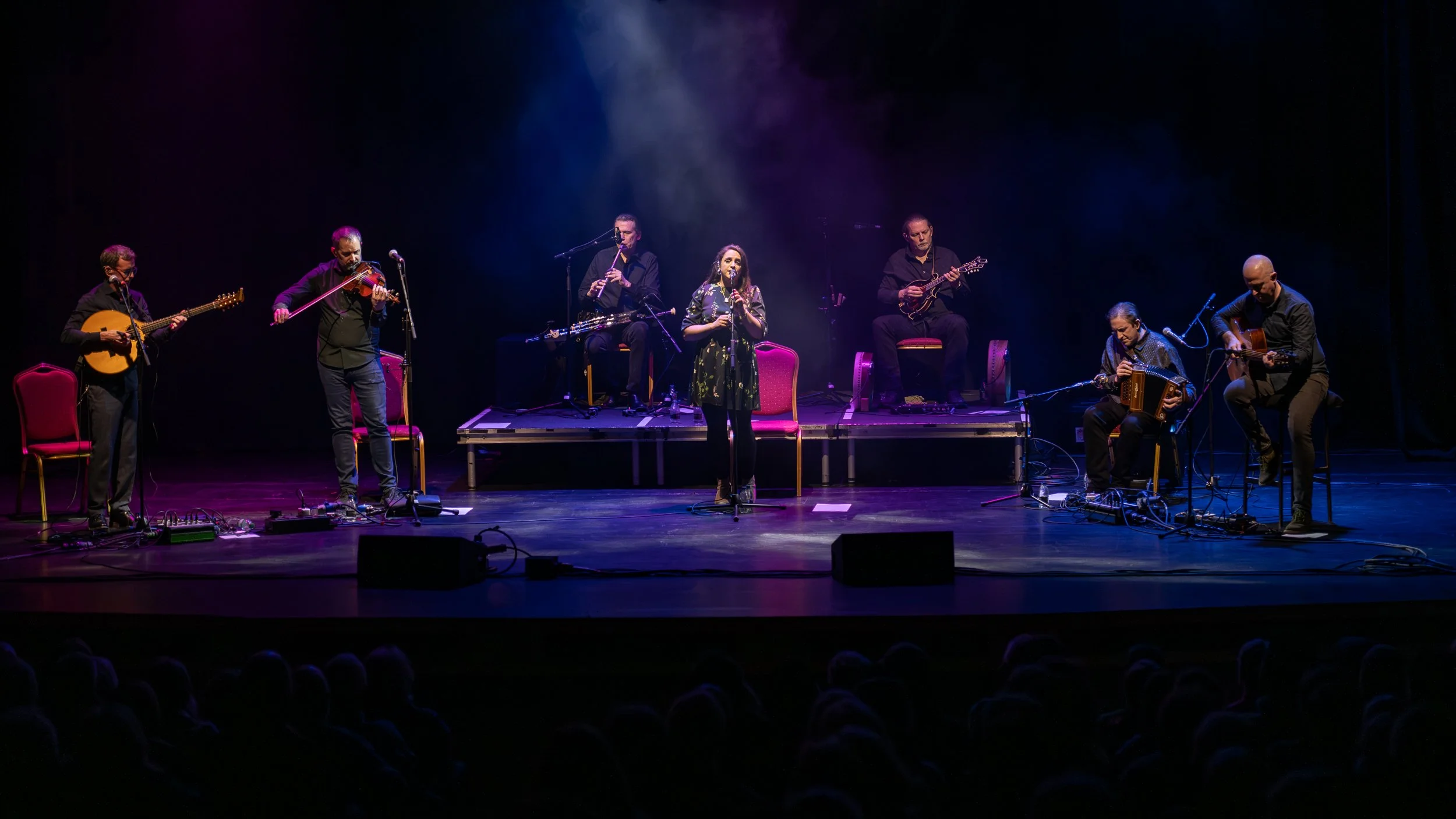 A band performing on stage with seven members, including a woman singing into a microphone, surrounded by several musicians playing guitars, a violin, a flute, and an accordion, with a dark background and stage lighting.