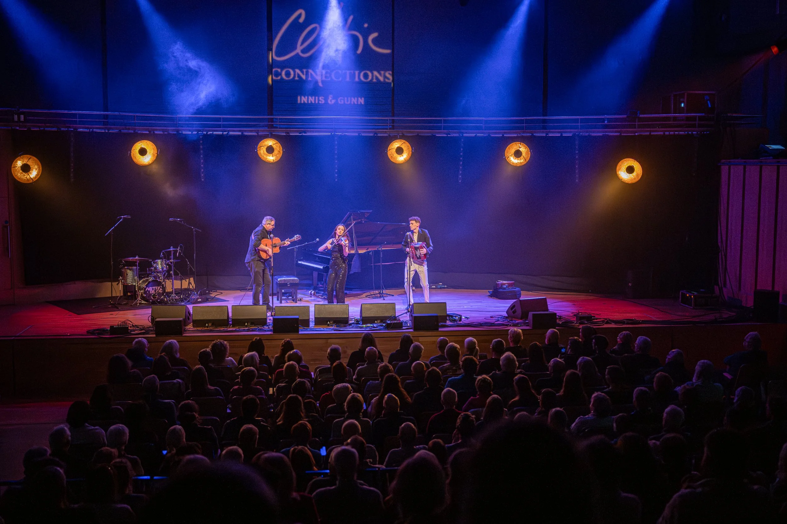 A musical performance on stage with three musicians, including a guitarist, a violinist and an accordion player, in front of an audience in a theatre with warm lighting.