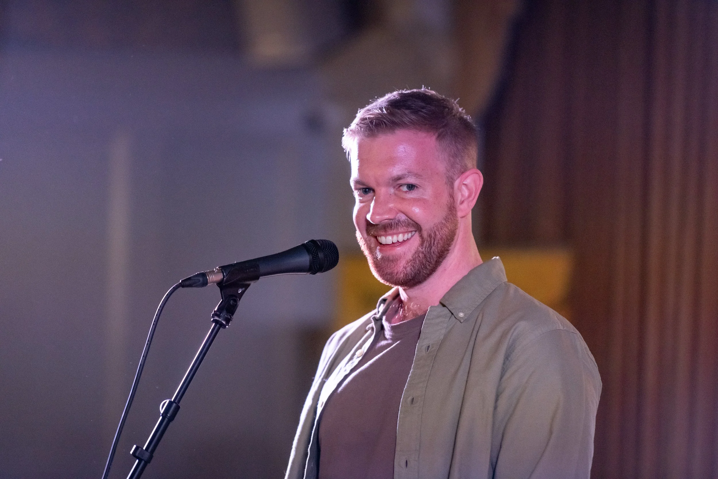 A smiling man with short brown hair and a beard, wearing a beige shirt, standing in front of a microphone on a stand with a blurred background.
